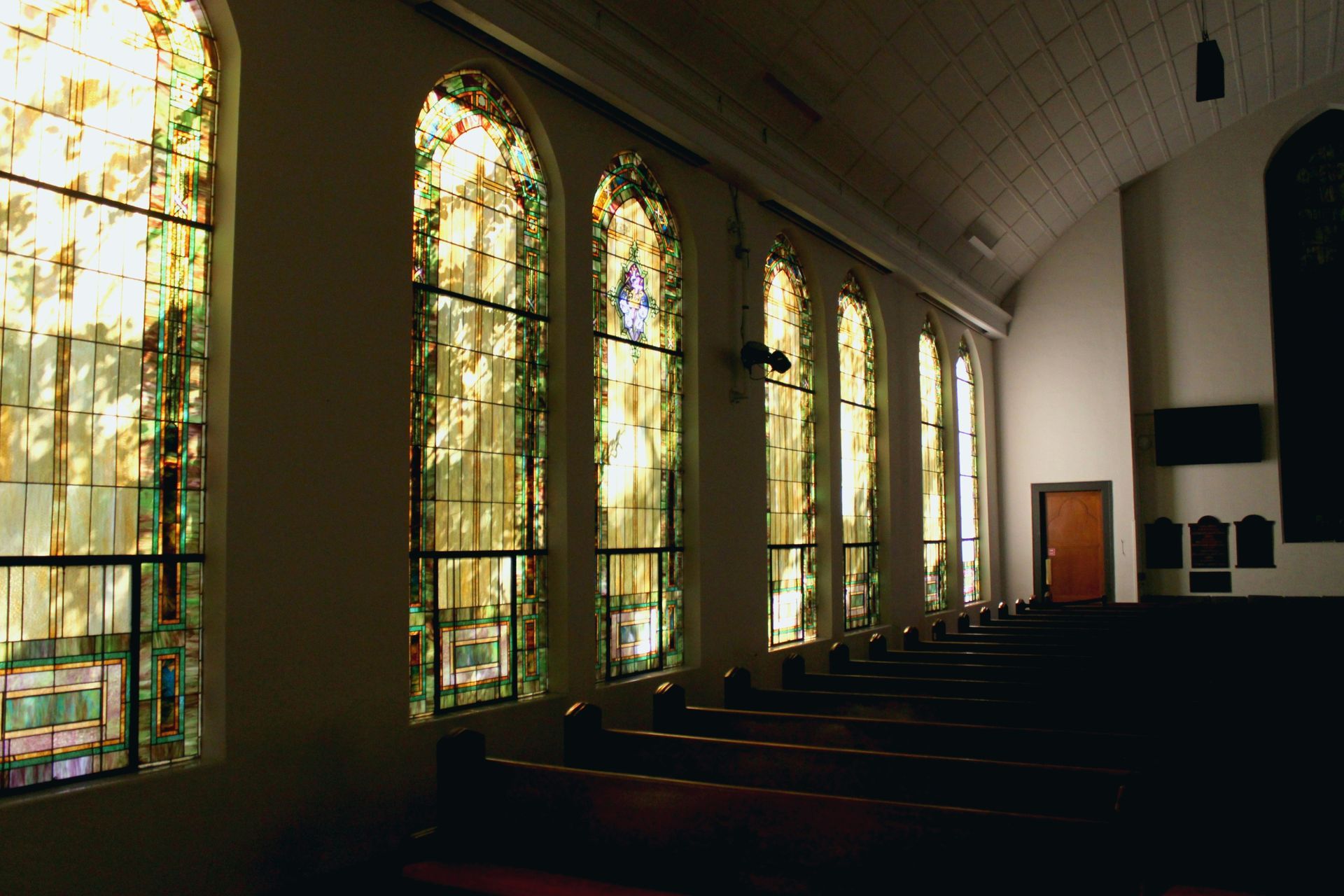 Interior of a church with rows of wooden pews facing an open doorway to a grassy area. Bright natural light.