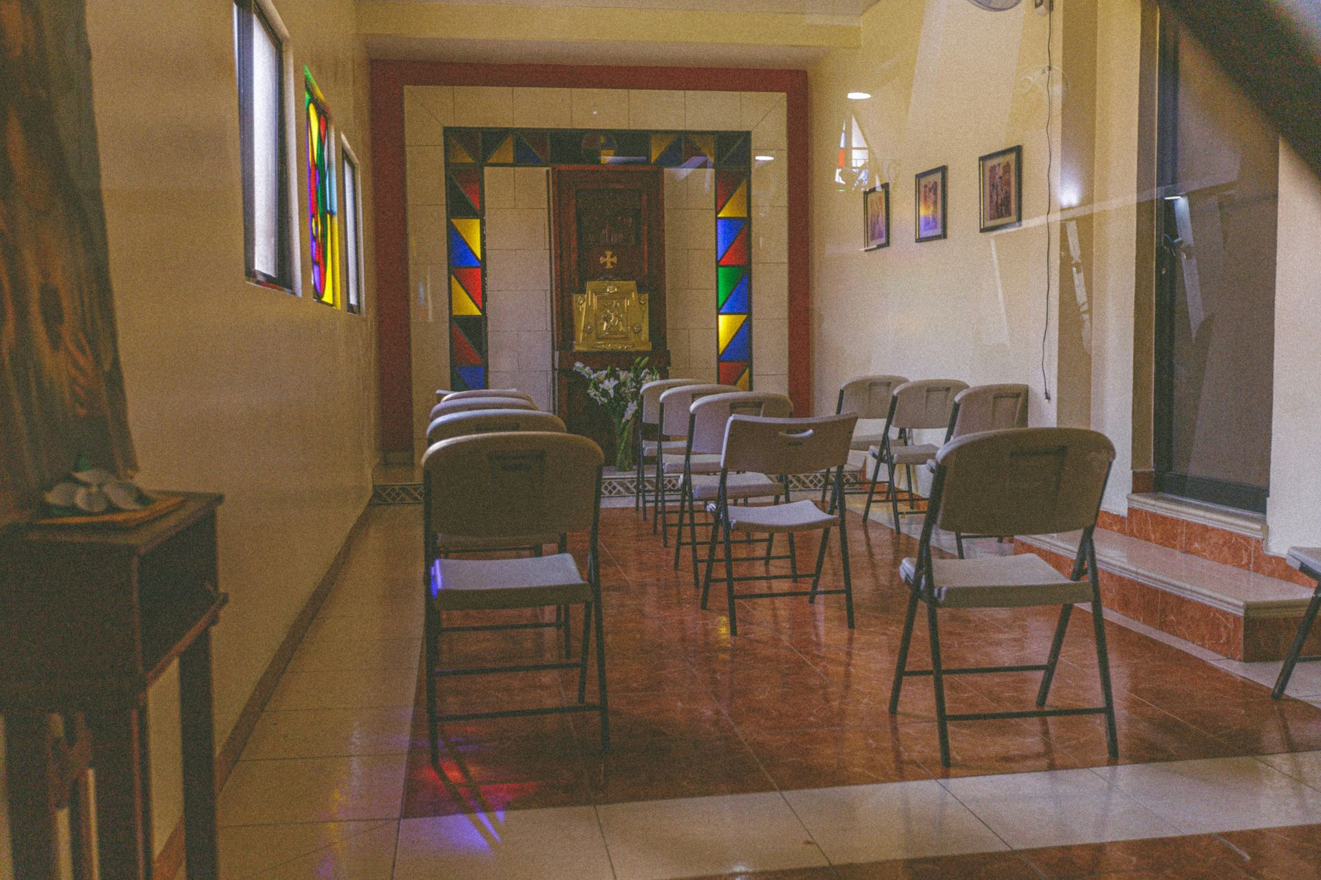 Small chapel with rows of chairs facing an ornate altar, colorful stained glass window, neutral walls.
