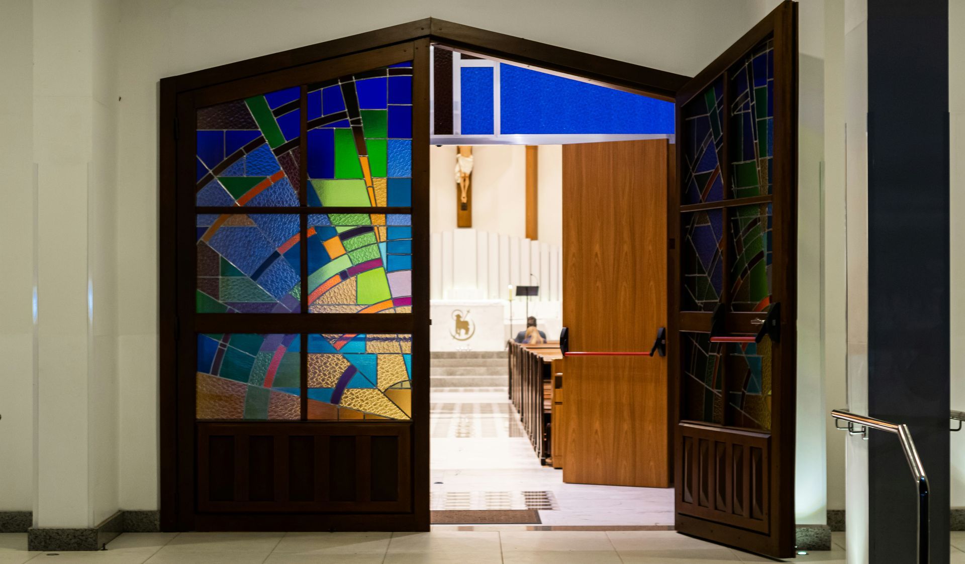 Interior of a church with rows of wooden pews facing an open doorway to a grassy area. Bright natural light.
