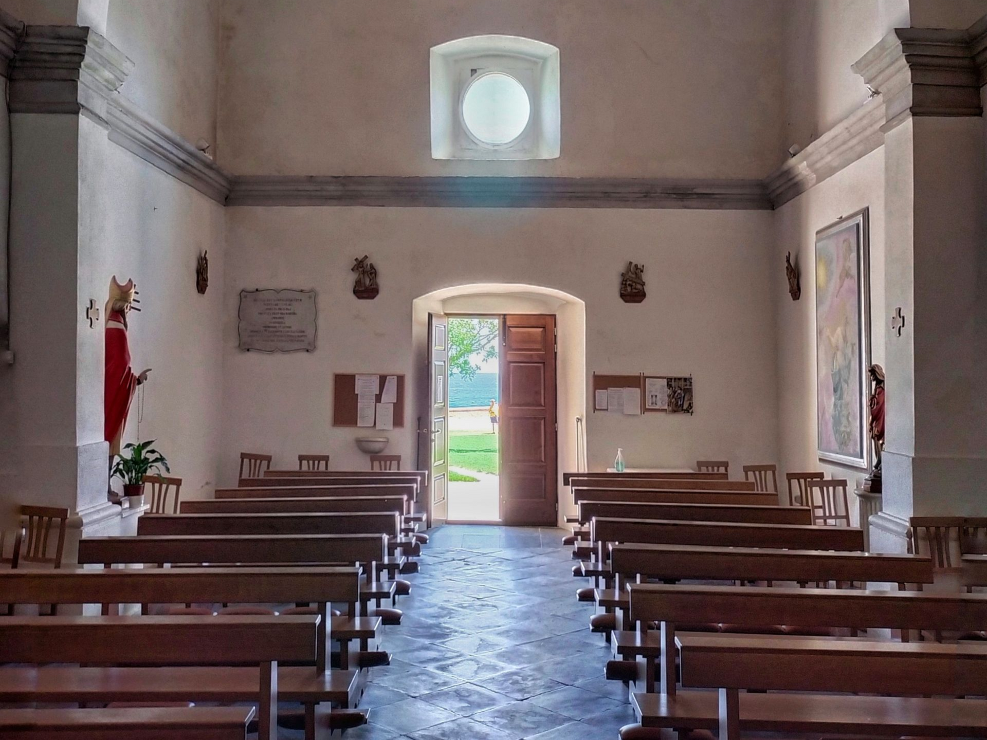 Interior of a church with rows of wooden pews facing an open doorway to a grassy area. Bright natural light.