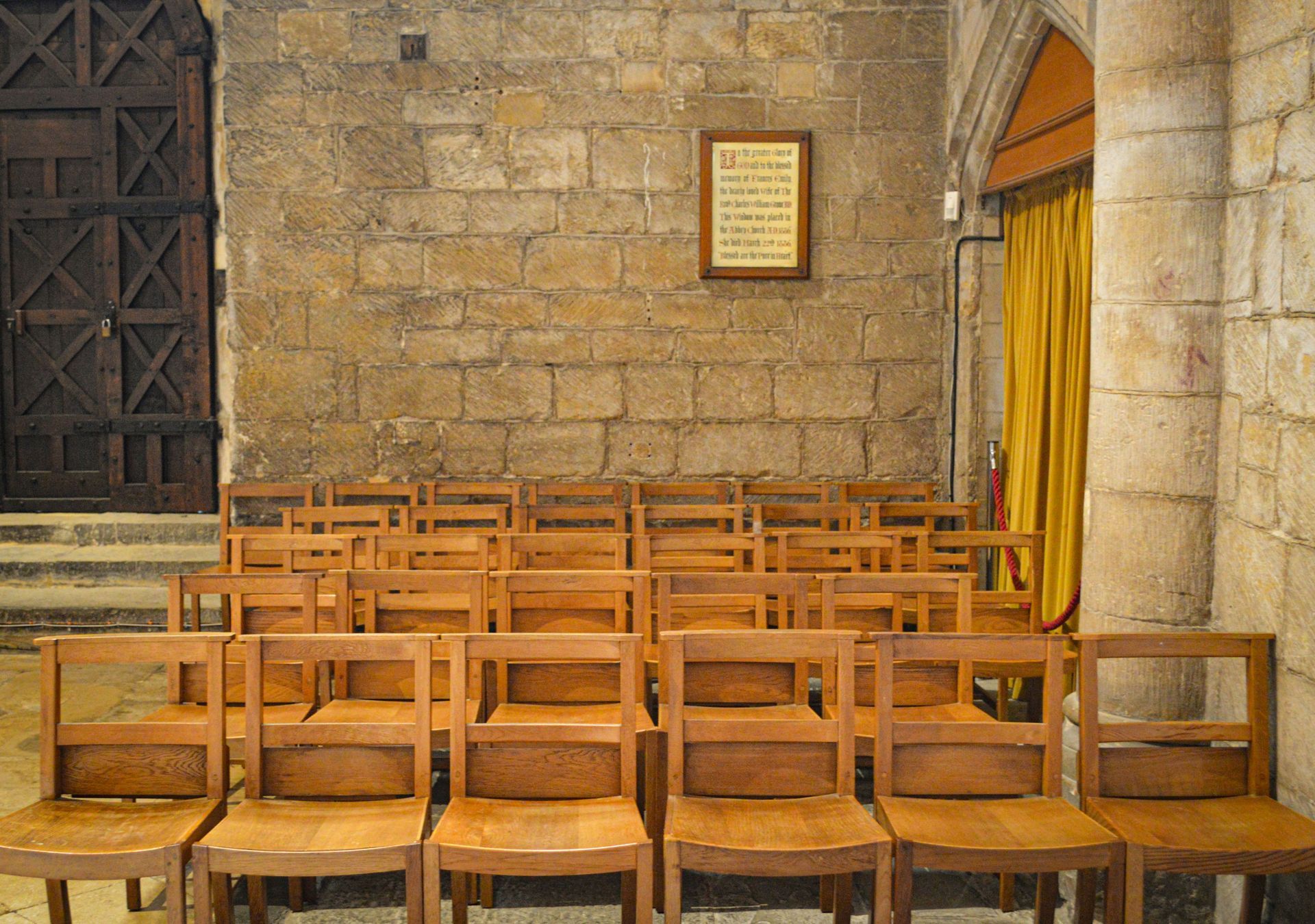 Rows of wooden chairs in a stone-walled room, likely a church. A sign hangs on the wall.