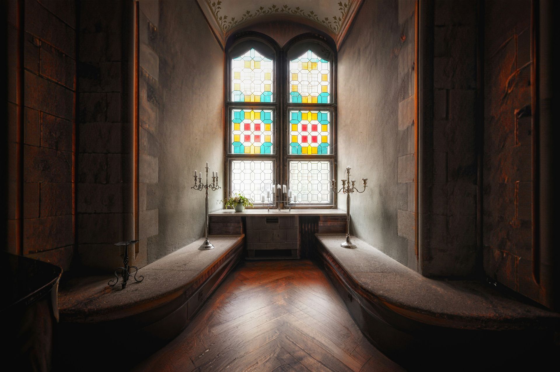 Narrow room with colorful stained glass window, benches, and candle holders.