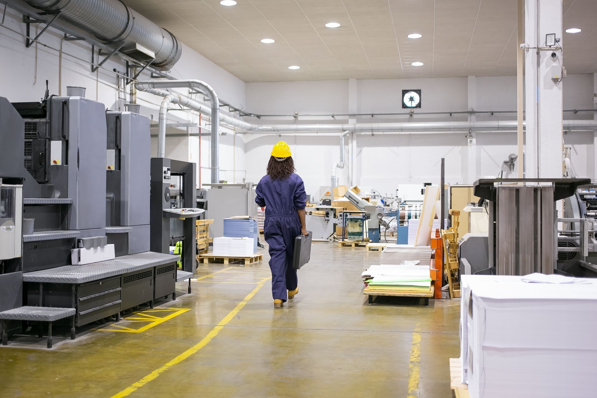 Woman in blue jumpsuit and yellow hard hat walks through a printing factory, carrying a case.
