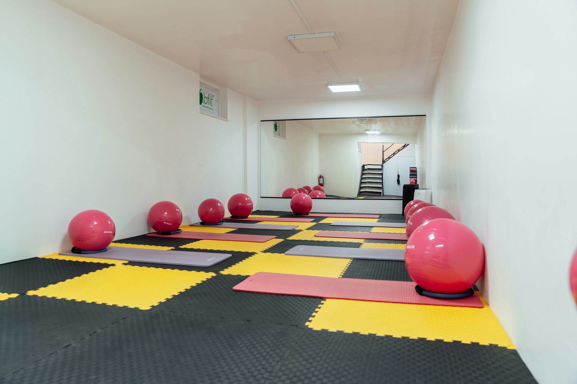 Yoga studio with pink exercise balls and mats on a black and yellow tiled floor, a large mirror reflects a staircase.