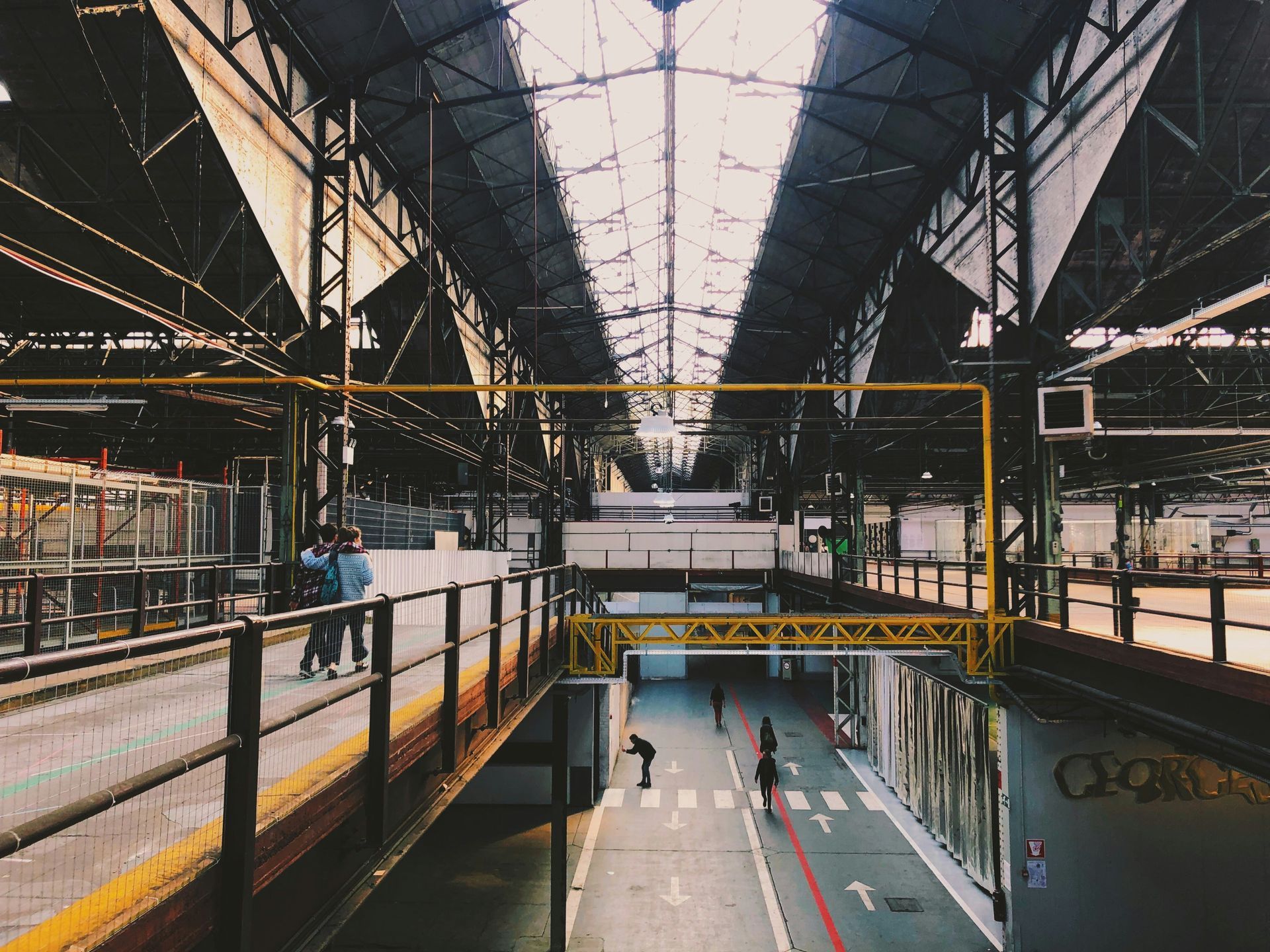 Inside a factory with metal roof, people walk on a raised walkway, with a road and more people below.