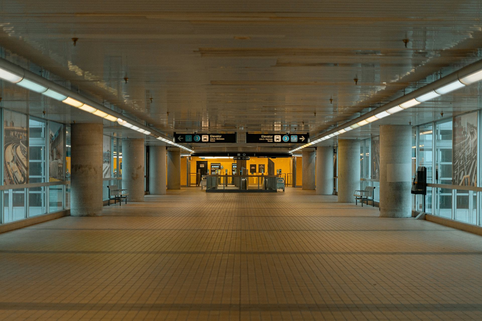 Interior of a subway station. Bright, long hallway with columns, lit by overhead lights, leading to turnstiles.