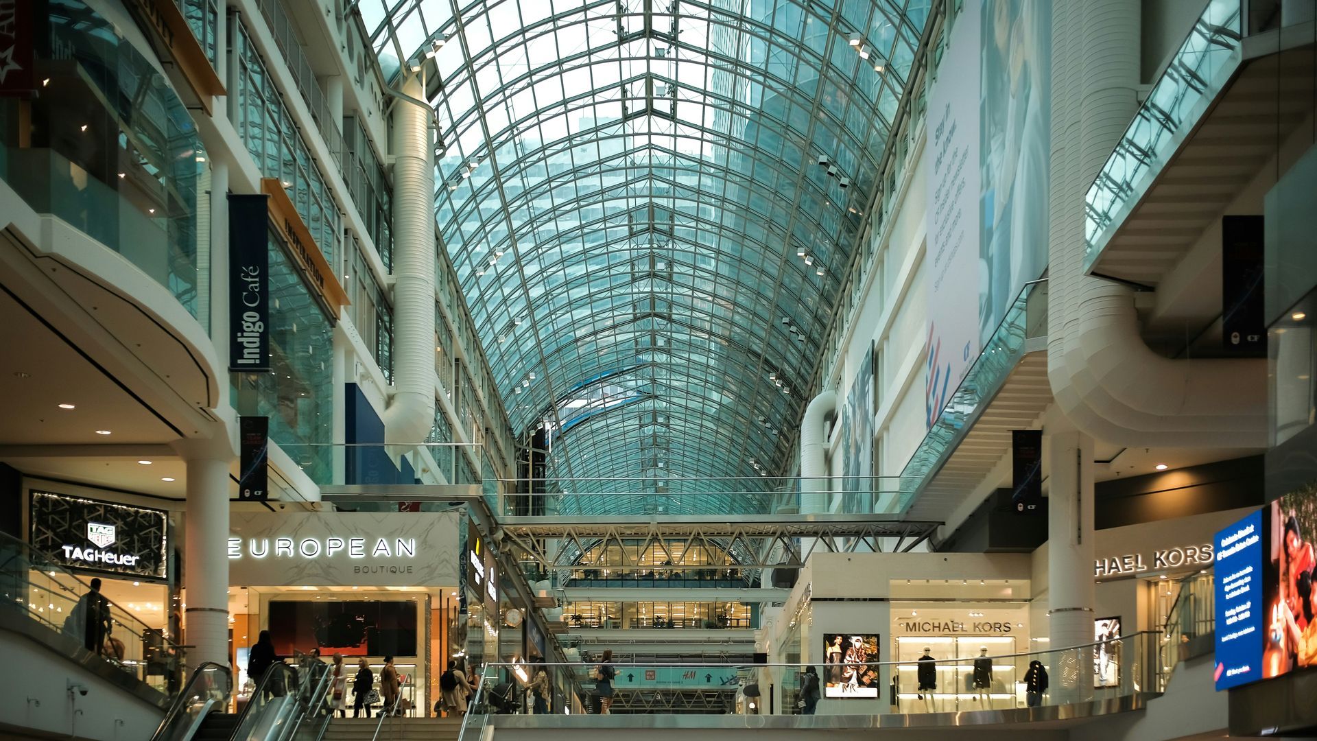 Interior of a large, upscale shopping mall with a glass ceiling and several stores visible.