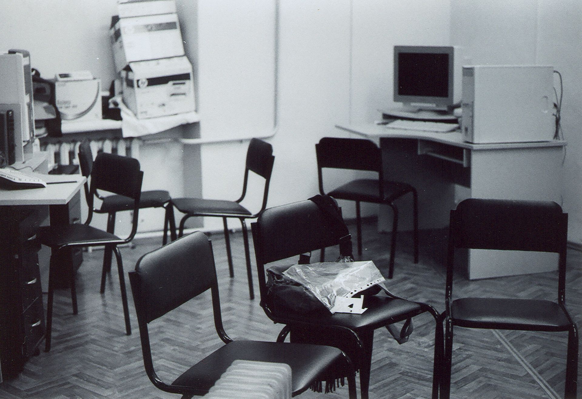 A cluttered office space with empty chairs, a desk with a computer, and boxes stacked in the corner.