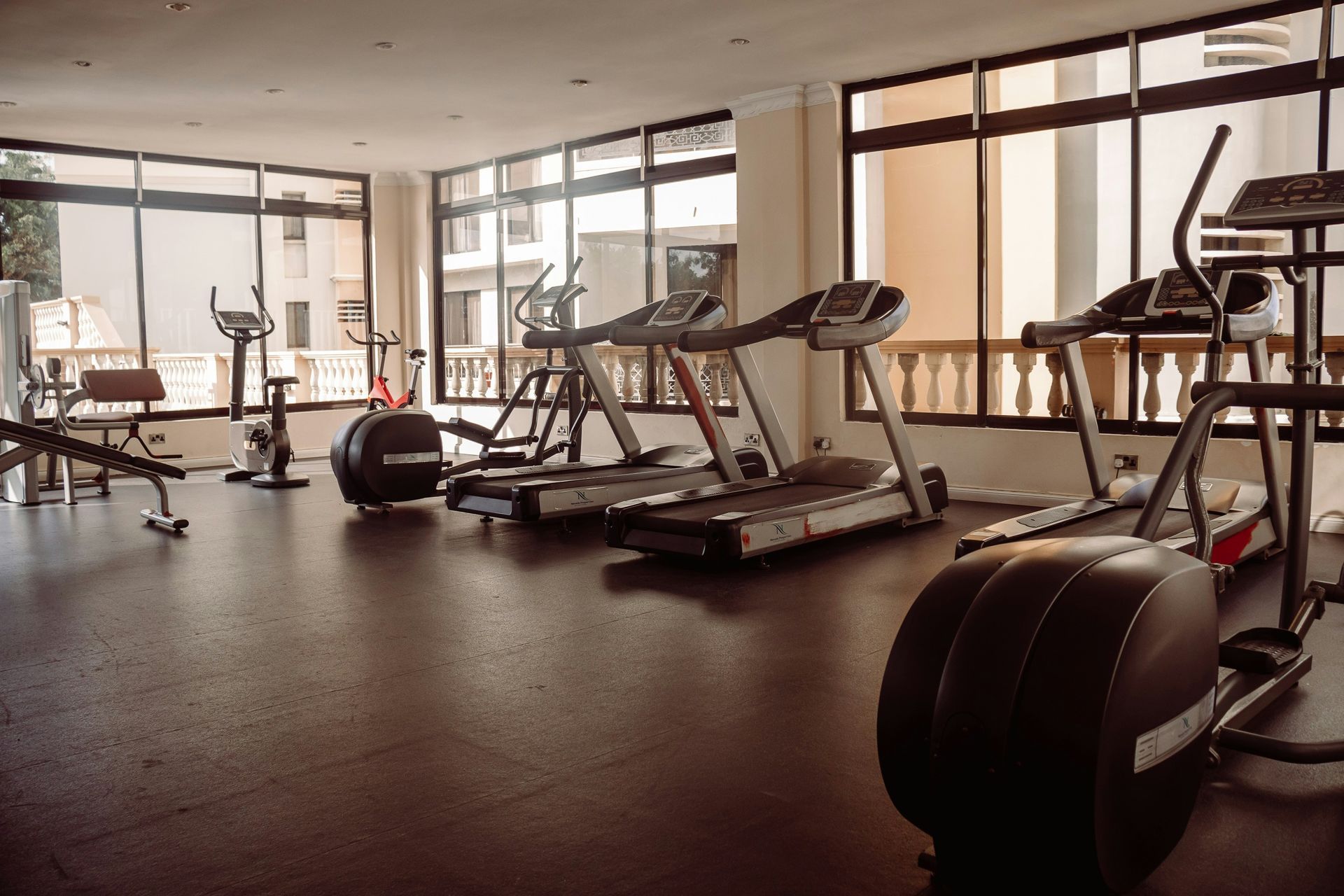 Gym interior with exercise equipment, including treadmills, stationary bikes, and weights, by large windows.