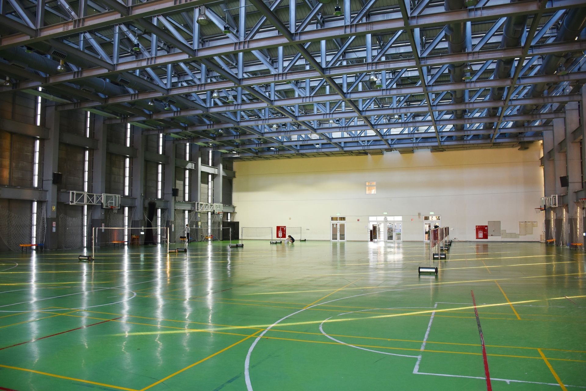 Indoor sports hall with green floor and metal beam ceiling; basketball hoops and badminton net visible.
