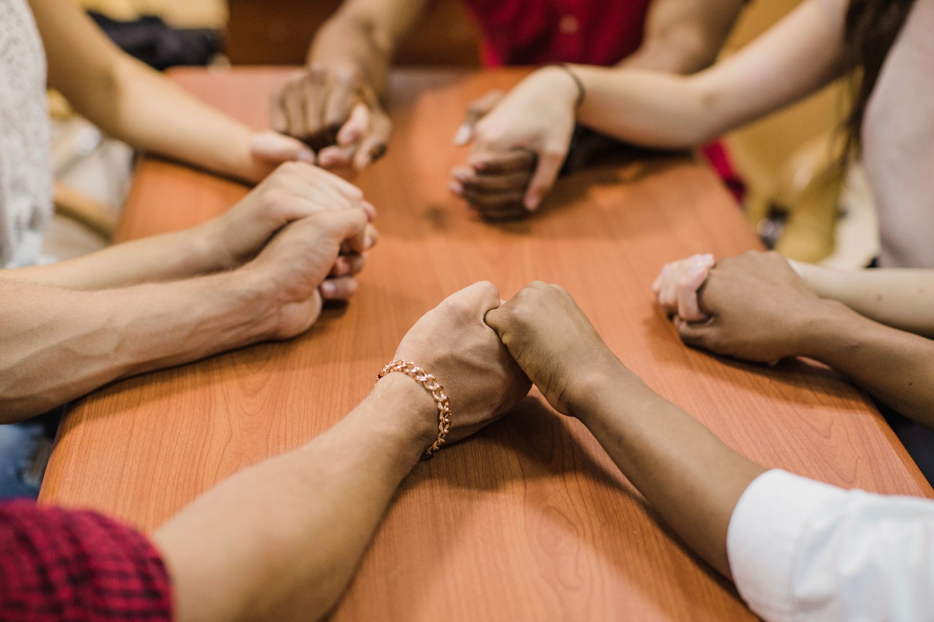 Hands clasped in prayer around a wooden table; diverse group.