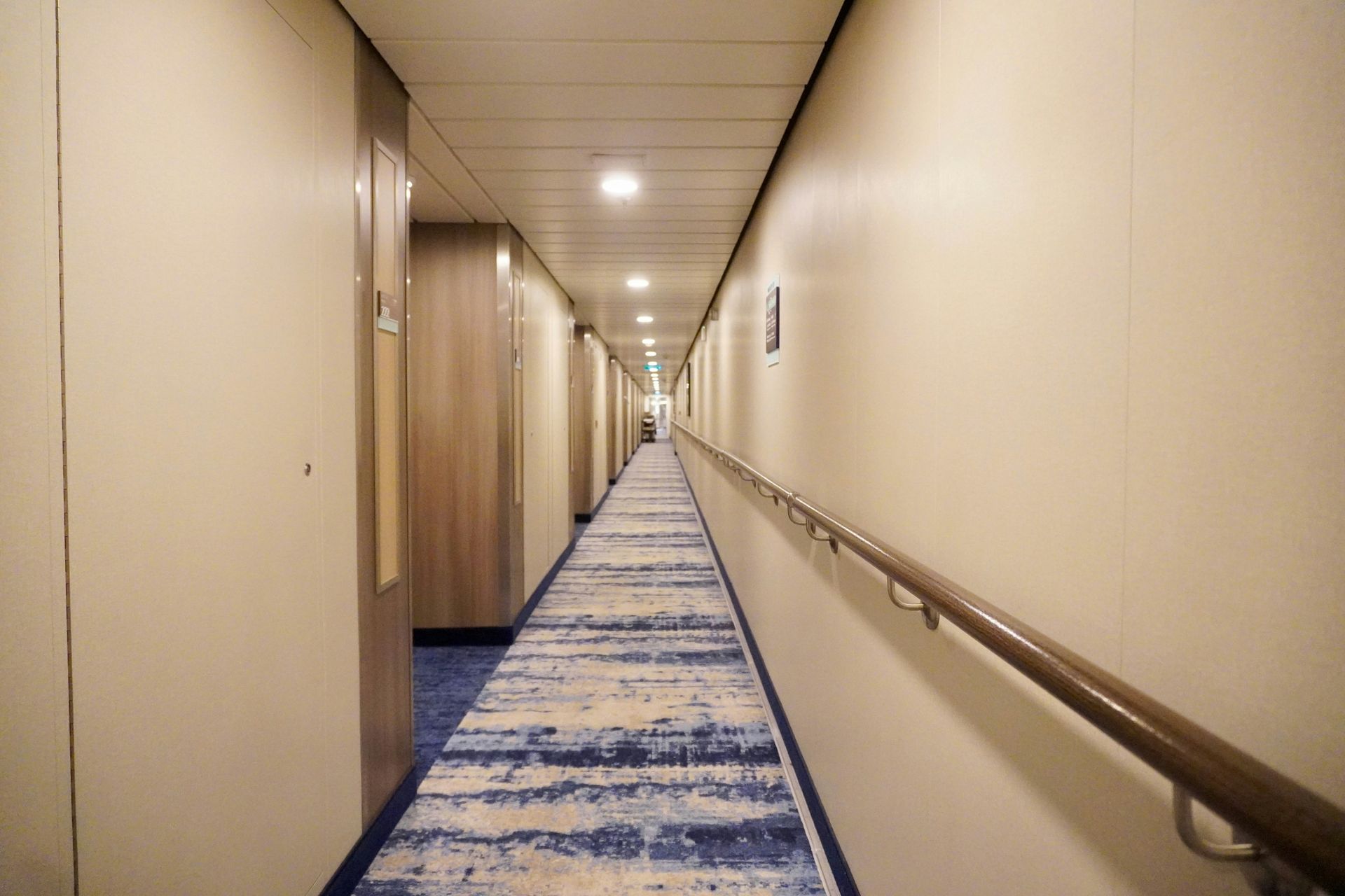 Long, narrow hallway on a cruise ship with blue carpet, cream walls, and doors on both sides.