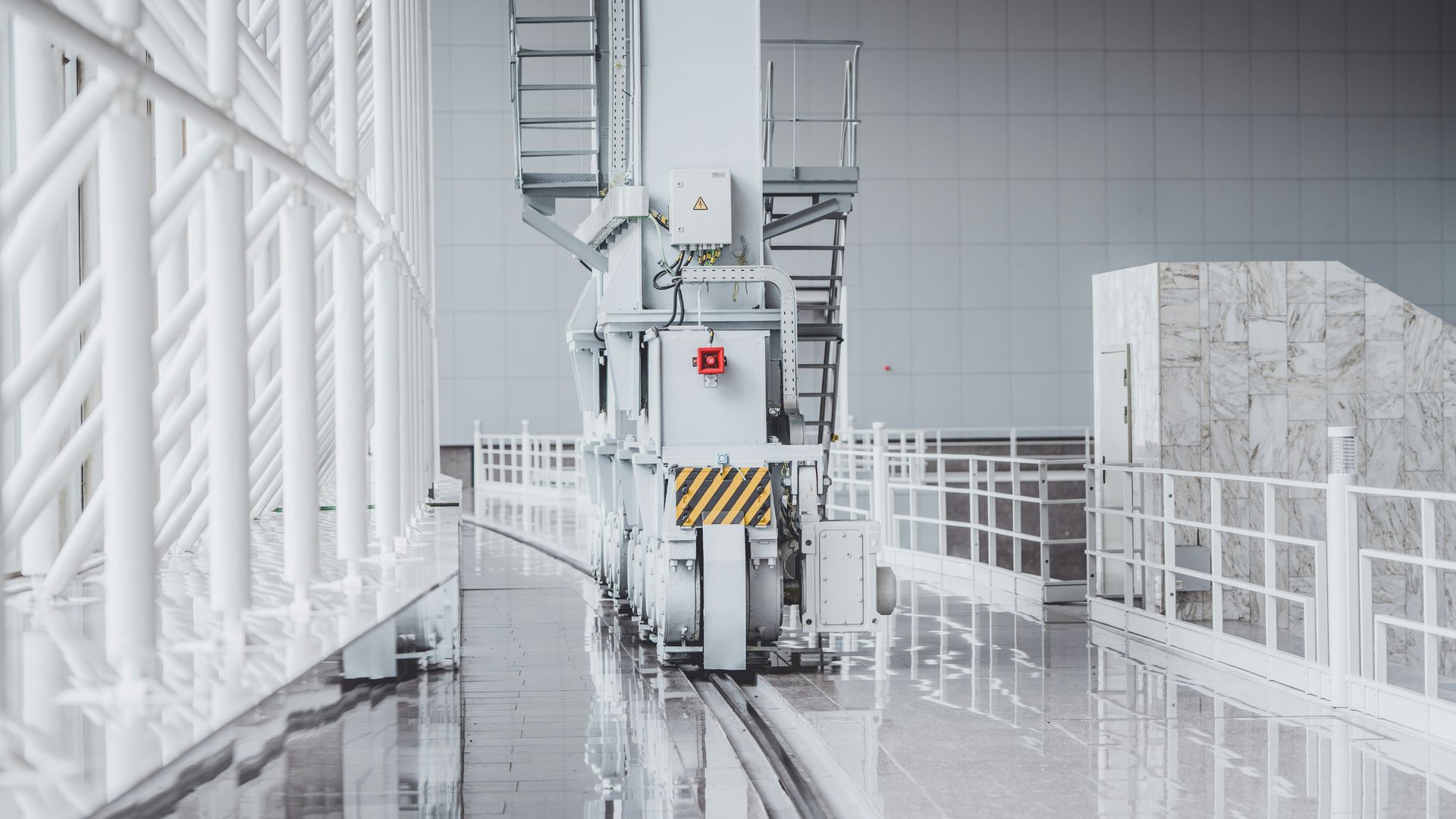 White industrial crane on tracks inside a building with a glossy floor and white railings.