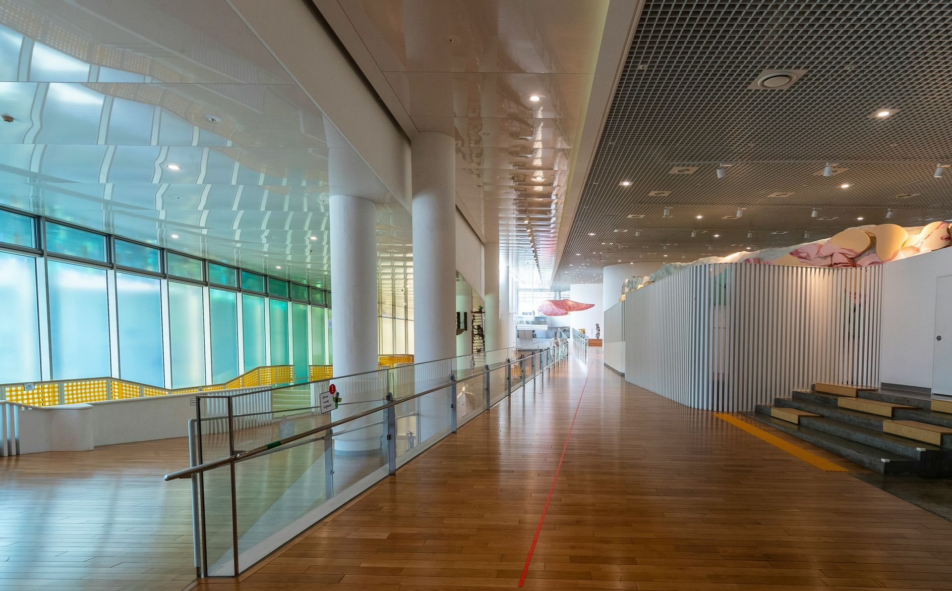 A long, bright museum hallway with a wood floor, columns, and colorful window light.