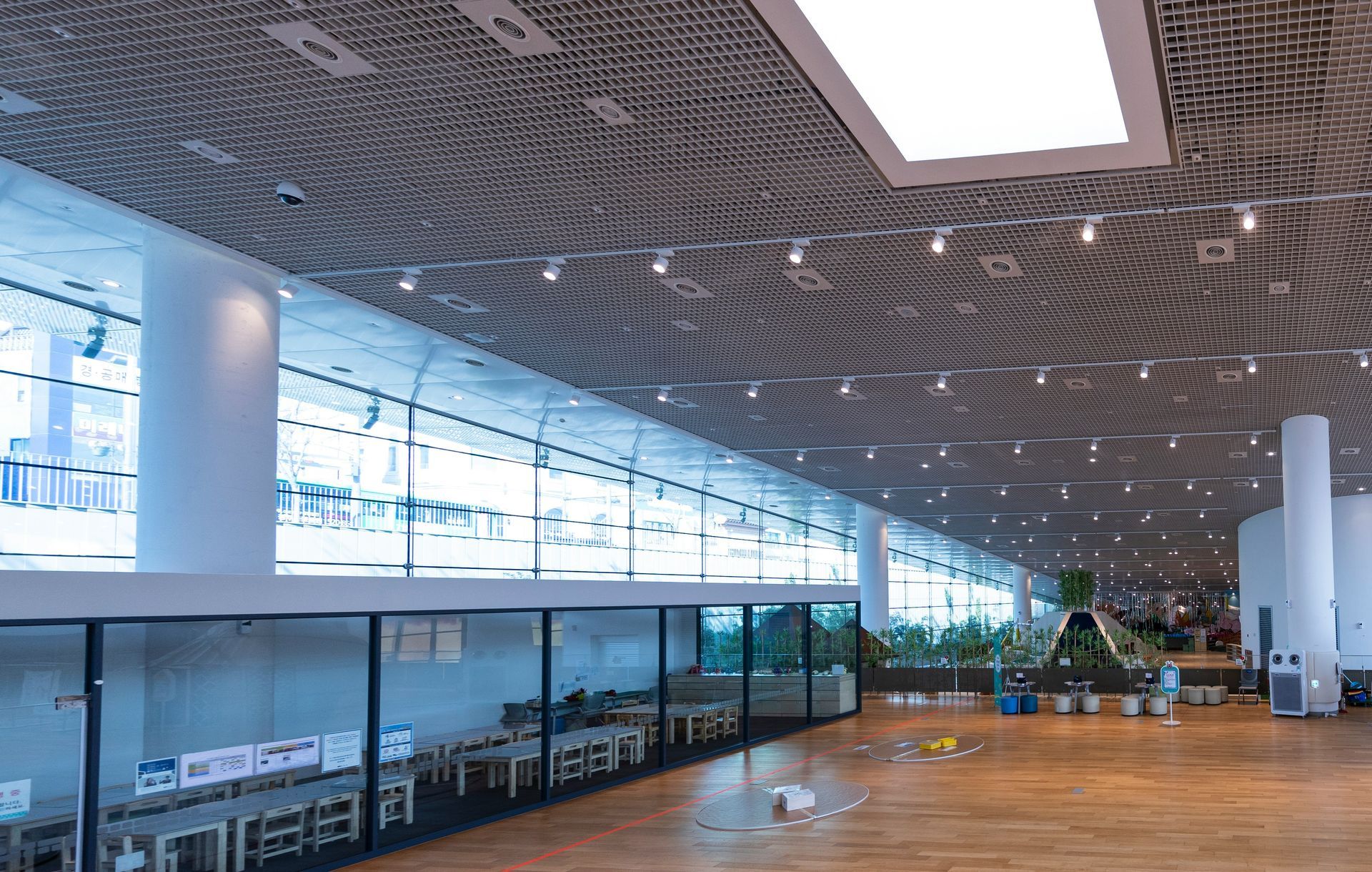 Spacious, modern interior with floor-to-ceiling windows, wooden floors, and long tables. White columns, patterned ceiling.