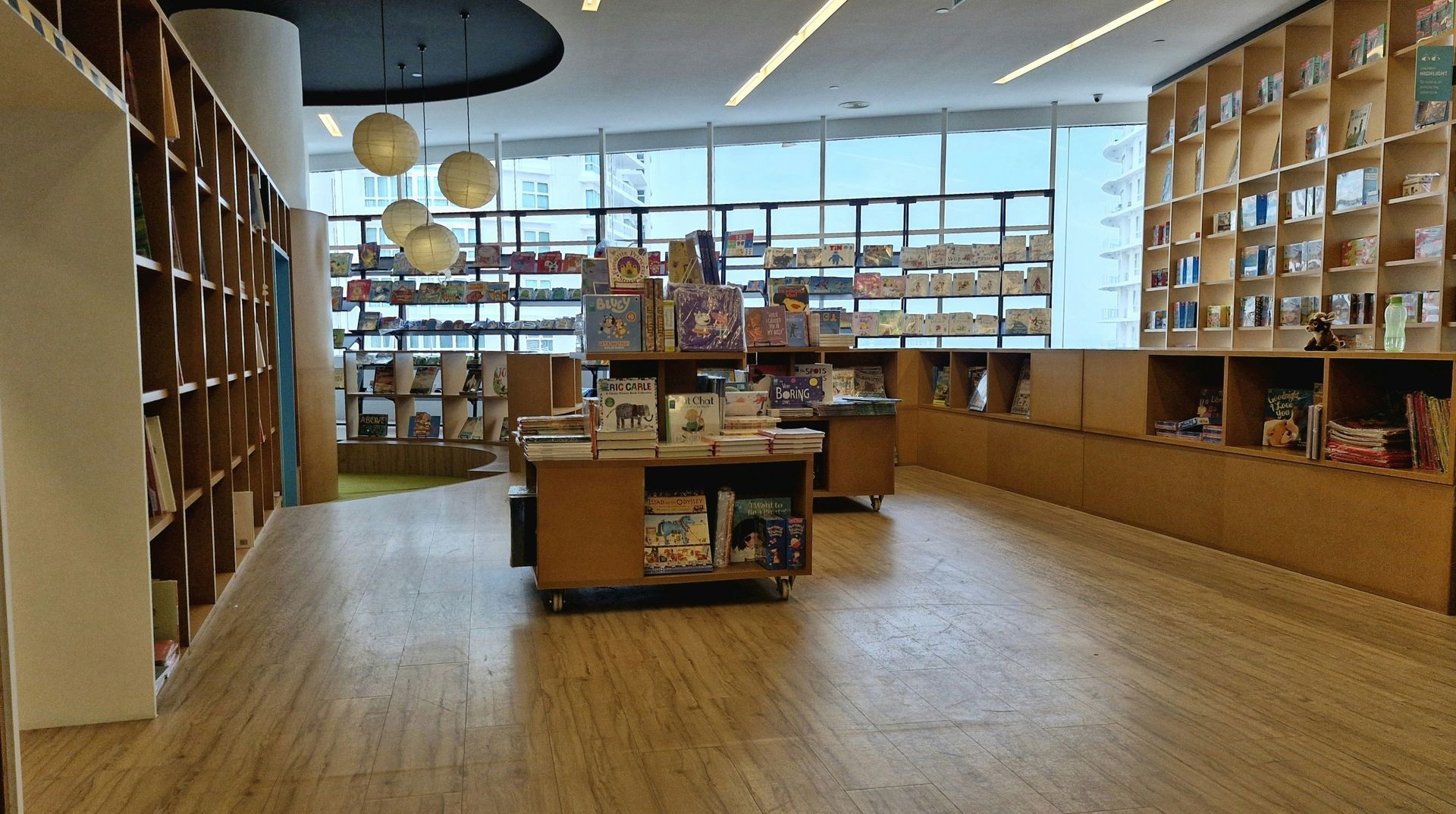 Interior view of a bookstore with wooden shelves and floors. Books are on display. Natural light from large windows.