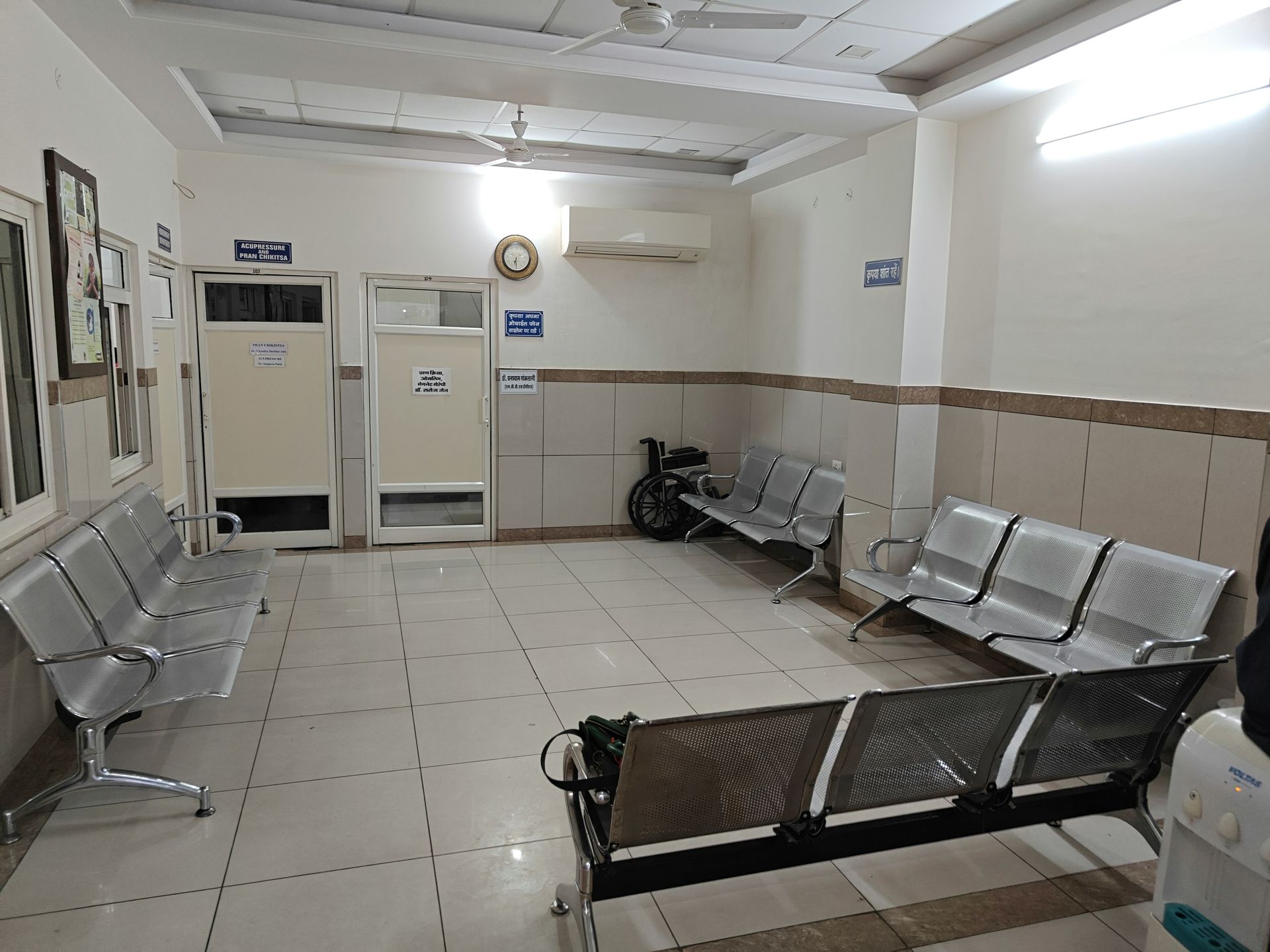 Waiting room with metal chairs, off-white walls, and doors. A wheelchair and air conditioner are visible.