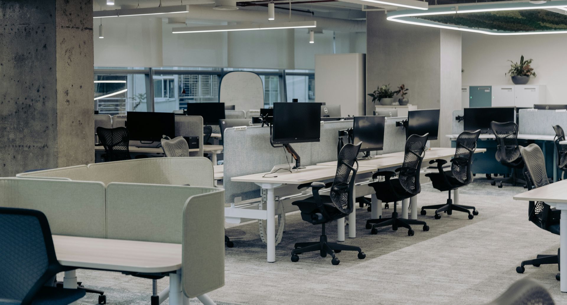 Modern office space with desks, computers, and chairs. Concrete column and light-colored partitions.
