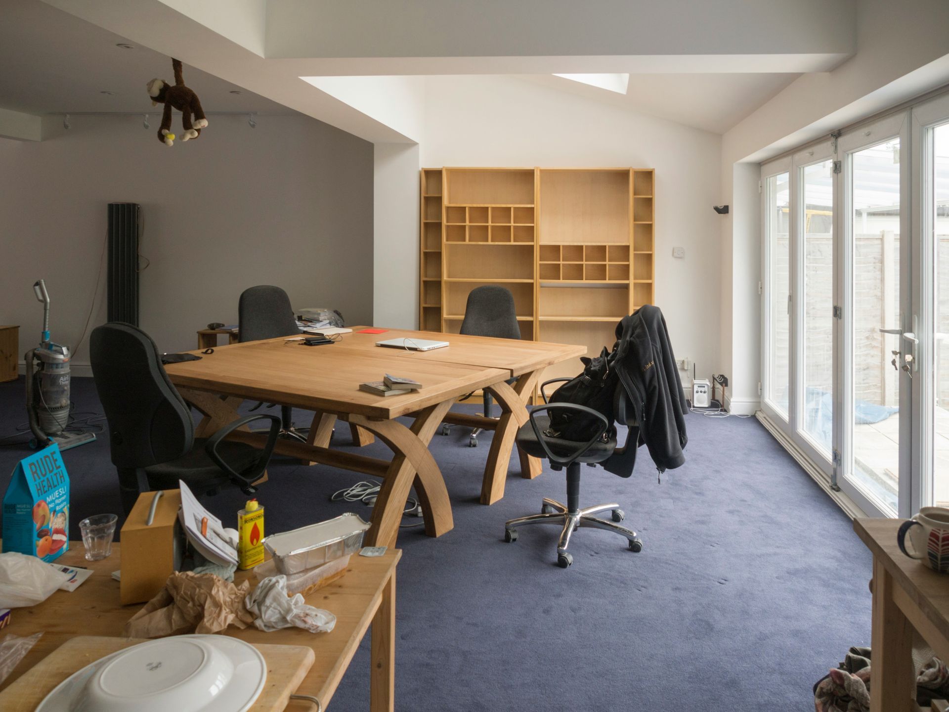 An office with a large wooden table and bookshelves. Windows on the right. Dark blue carpet and cluttered.