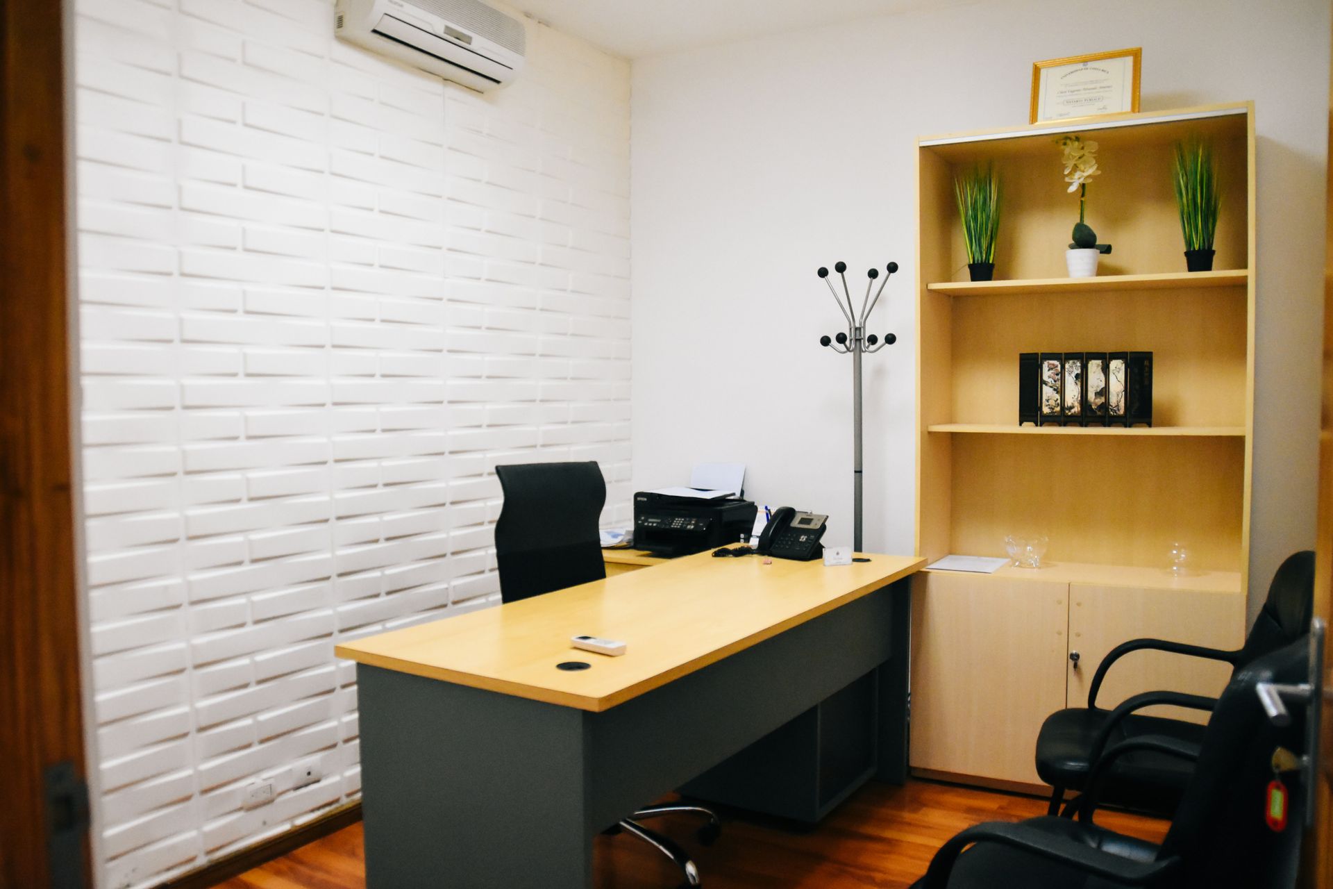 Office with desk, shelves, plants, and two black chairs. White brick wall and wooden floors.