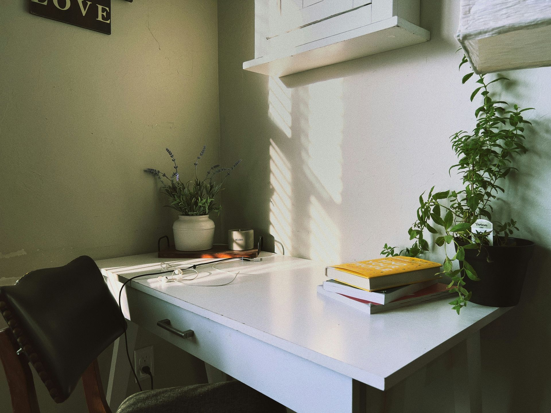 White desk with books, plants, and a chair in a sunlit corner.