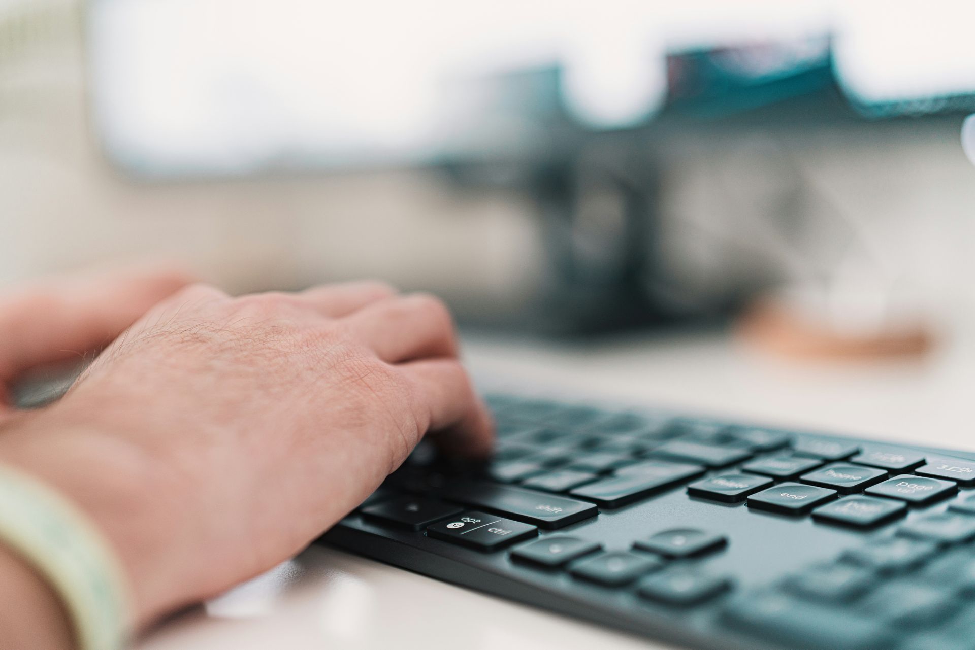 Hands typing on a black keyboard with a blurred computer monitor in the background.