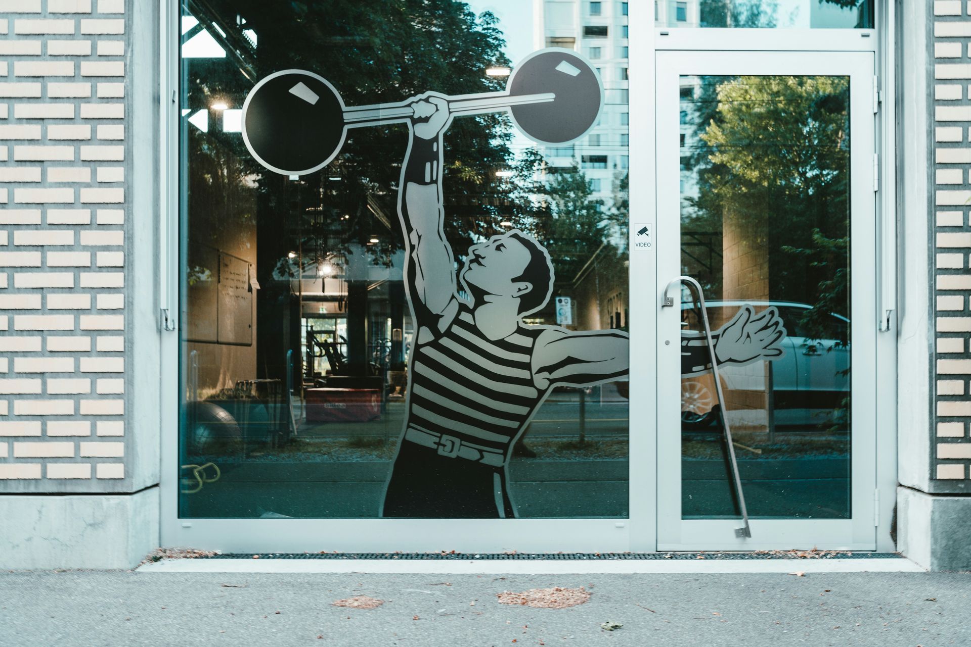 A glass door with a weightlifter graphic, reflecting a street scene.