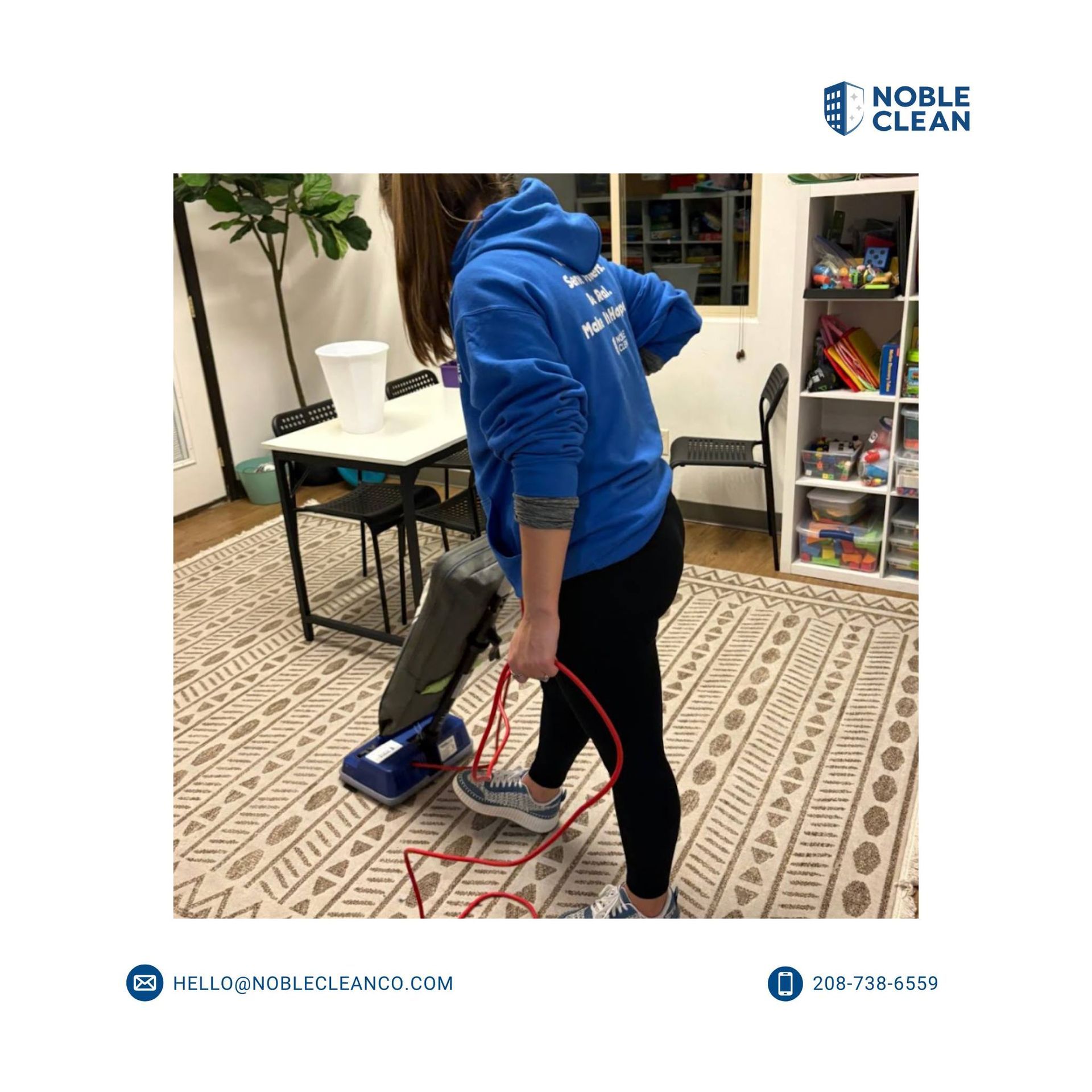 A person in a blue hoodie vacuums a patterned rug in a bright room with a table, chair, and storage shelves.