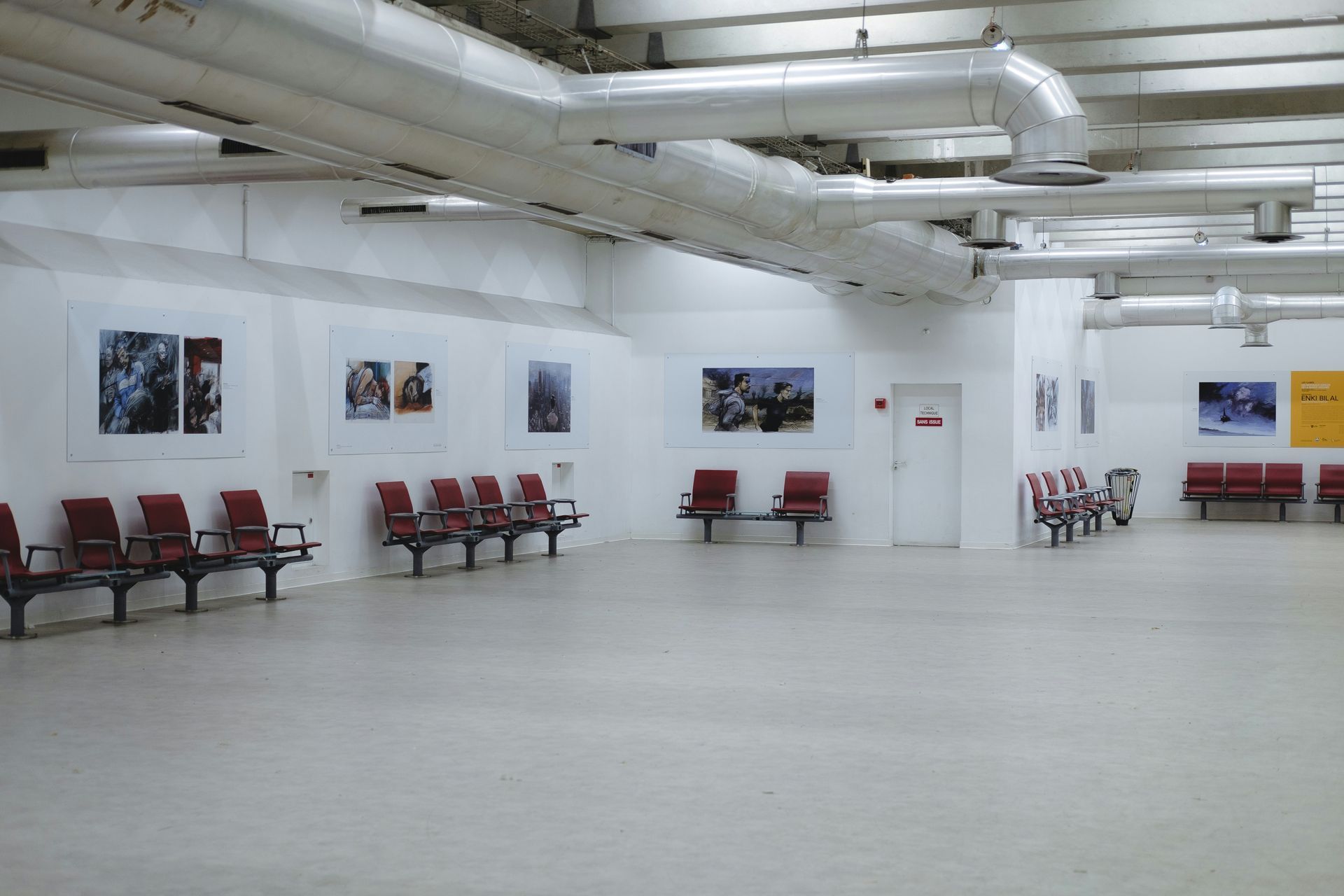 Empty airport waiting area with rows of red seating and artwork on the walls.