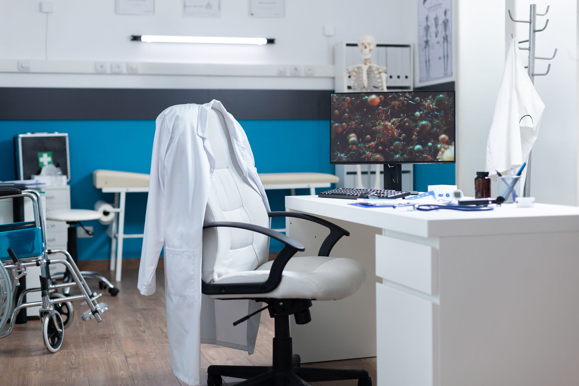 Doctor's office with a white coat on chair, desk with medical tools, wheelchair, and medical equipment.