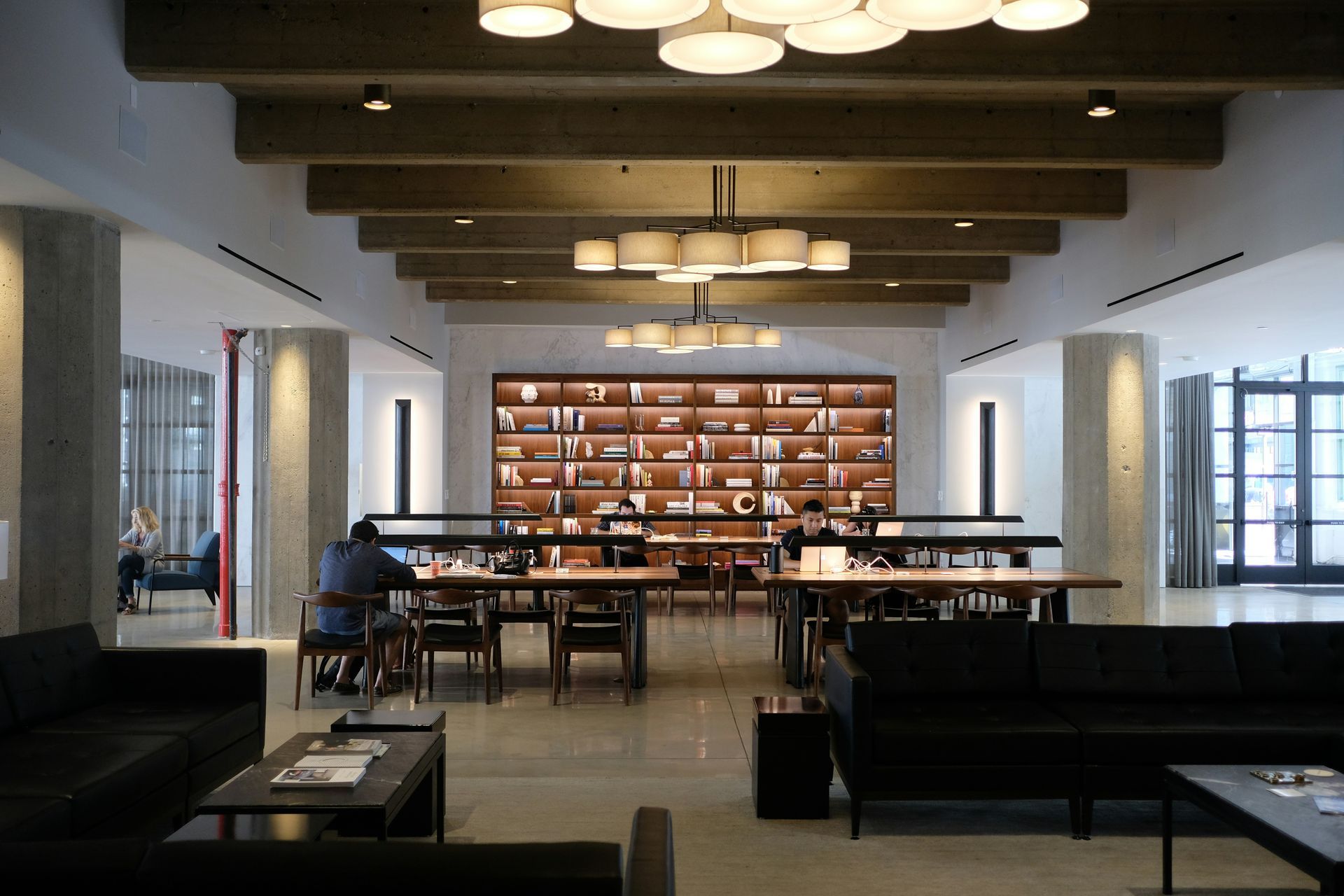 A modern library interior with wood tables, chairs, sofas, and a large bookshelf, with people reading.