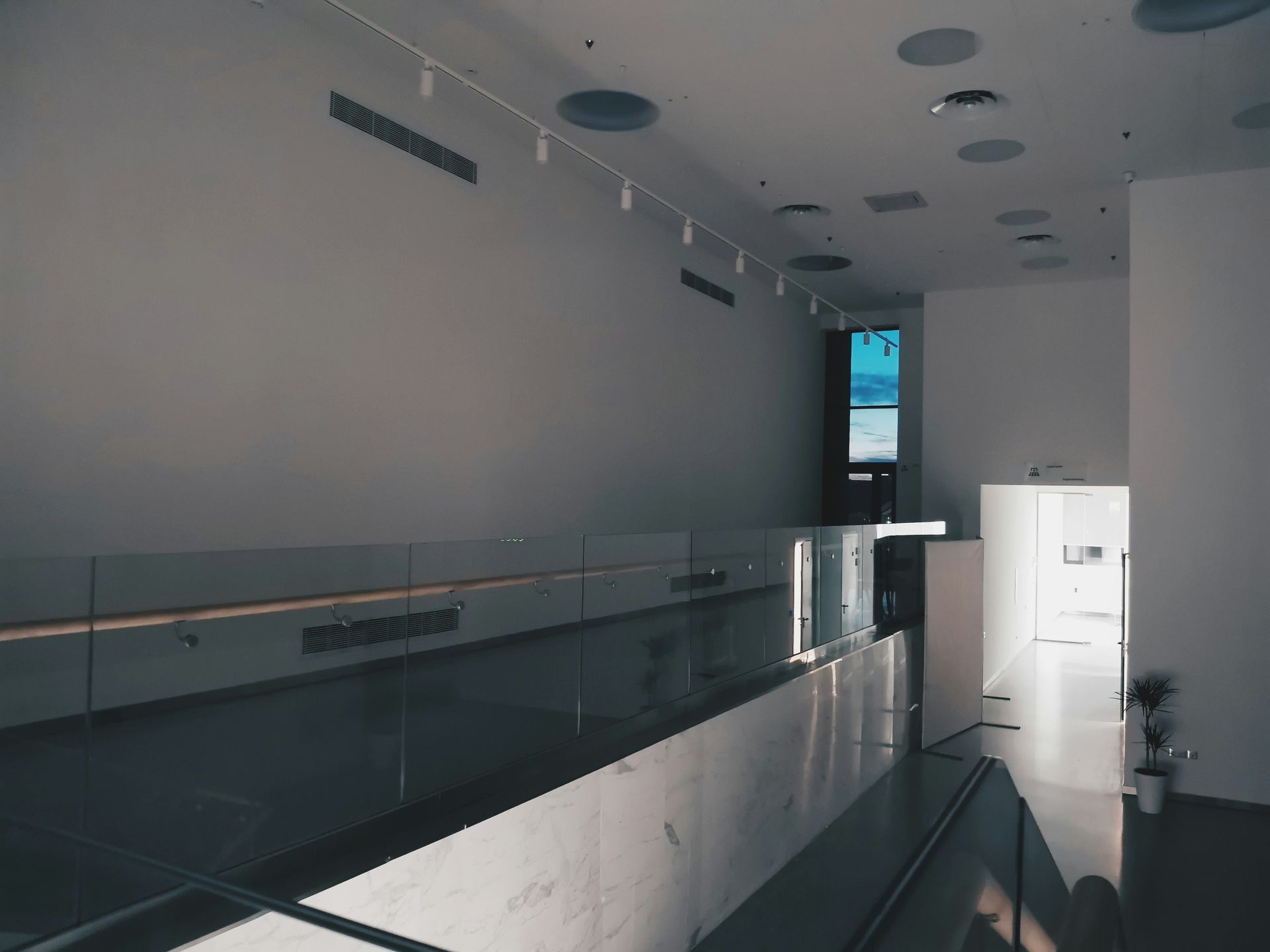 Long, empty gallery with white walls, glass railings, and a distant window showing blue sky.