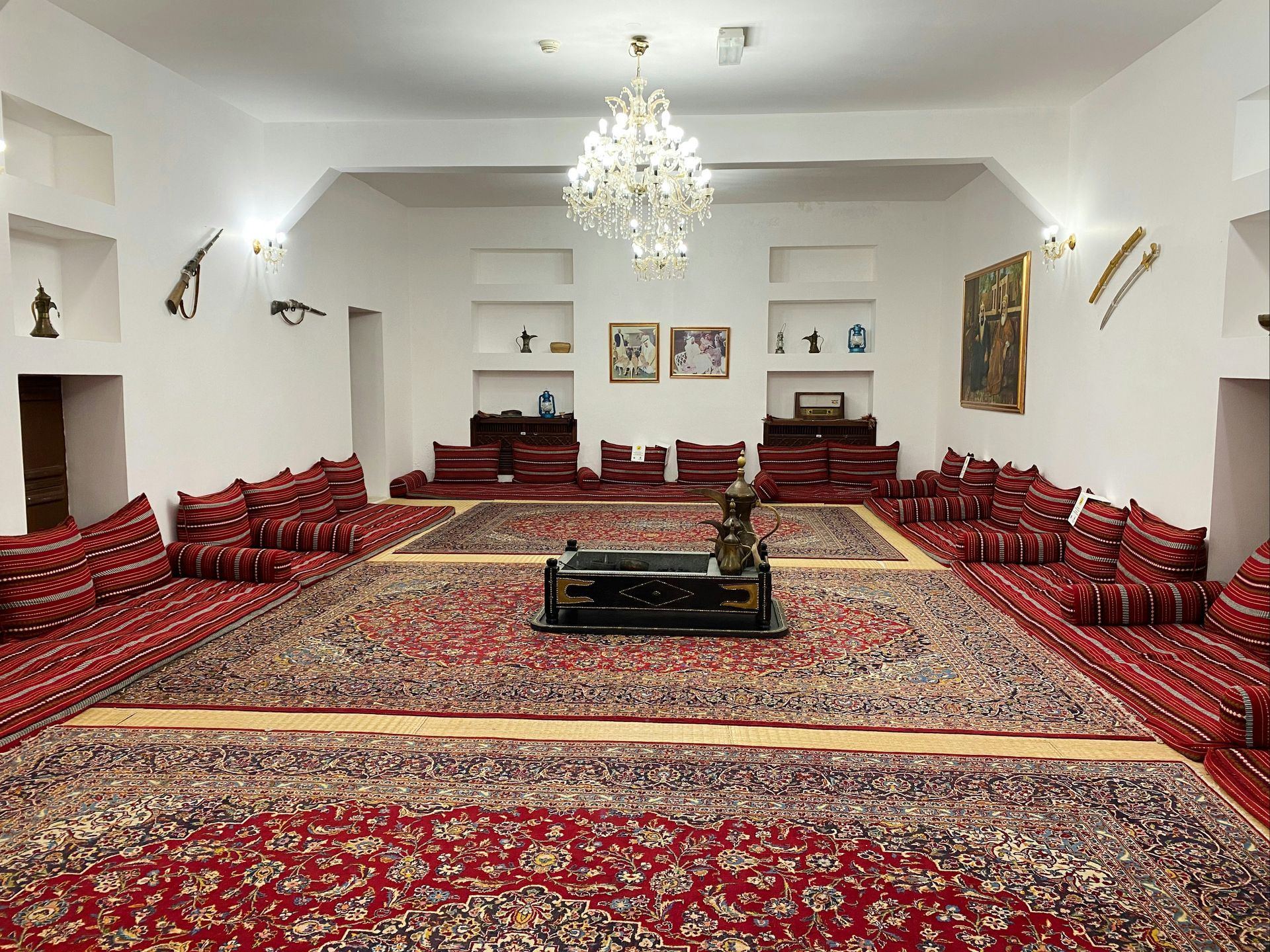 A room with red carpets, cushions, and traditional Middle Eastern decor; chandelier overhead.