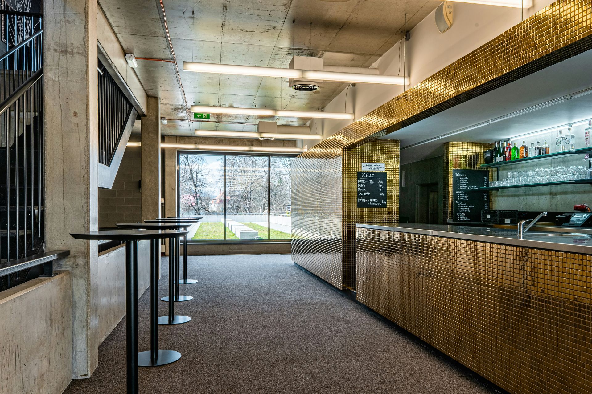 Bar area with gold facade, tables, window overlooking a green space, and a concrete staircase.