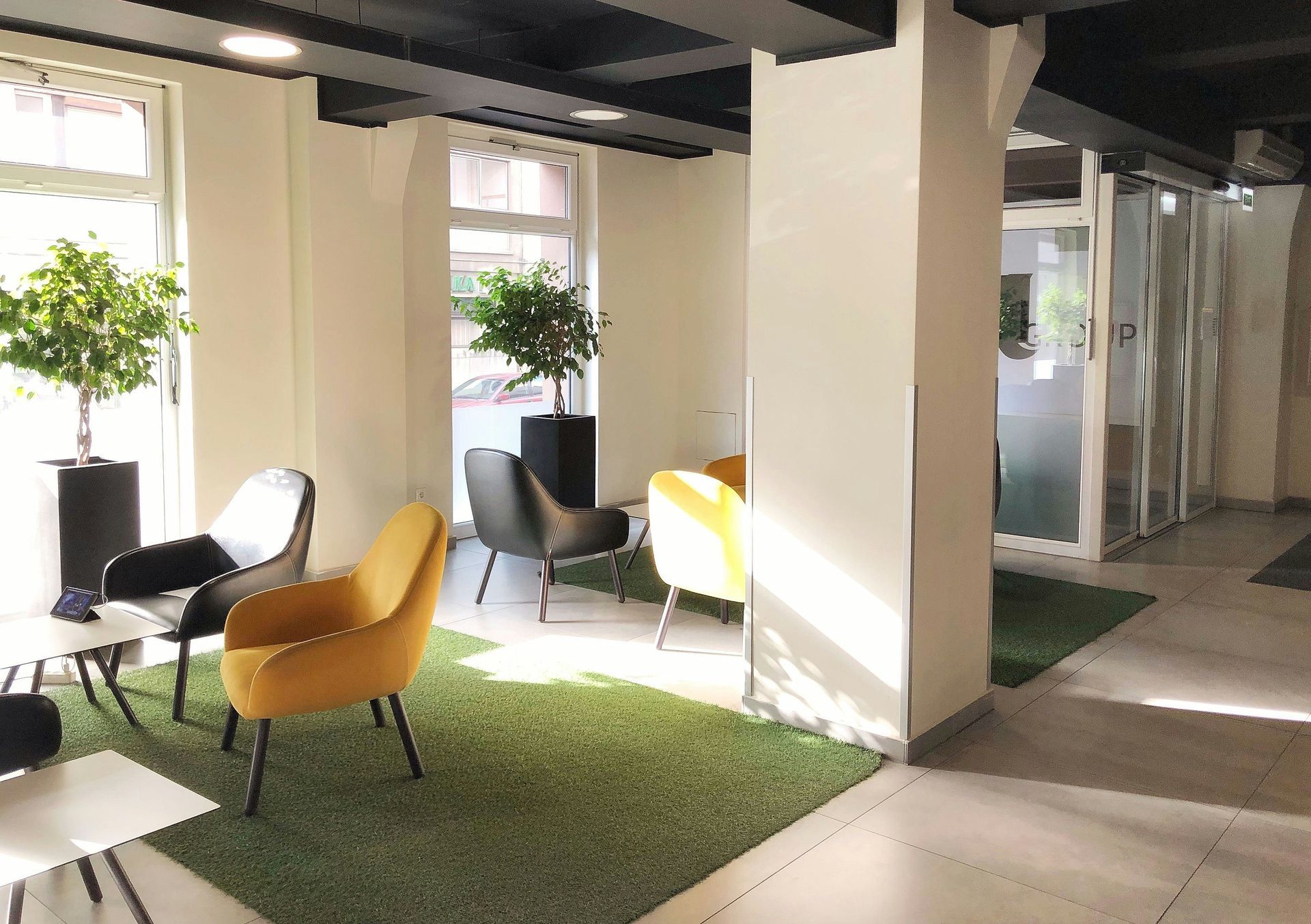 Lounge area with yellow and black chairs, artificial grass, and potted trees. Bright sunlight enters the room.