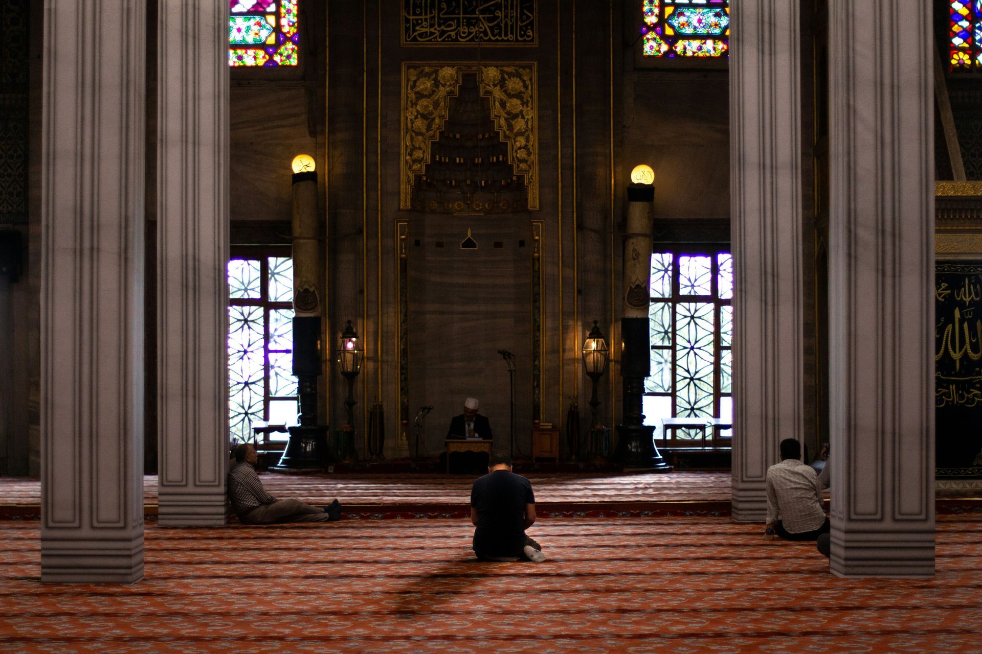 People praying in a mosque with large columns and stained glass windows; a red carpet.