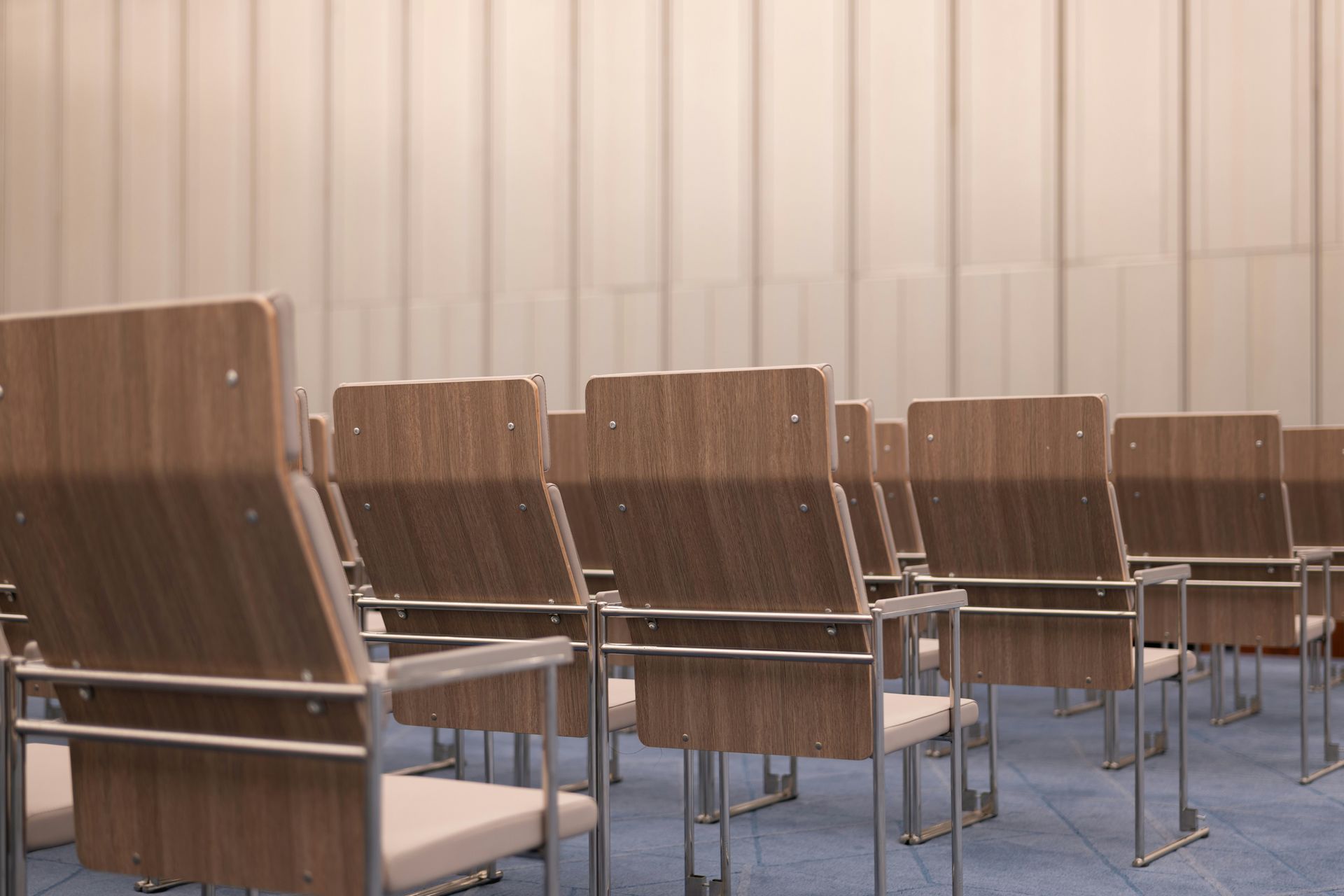 Rows of empty wooden chairs with metal frames in a conference room, facing a neutral-colored wall.
