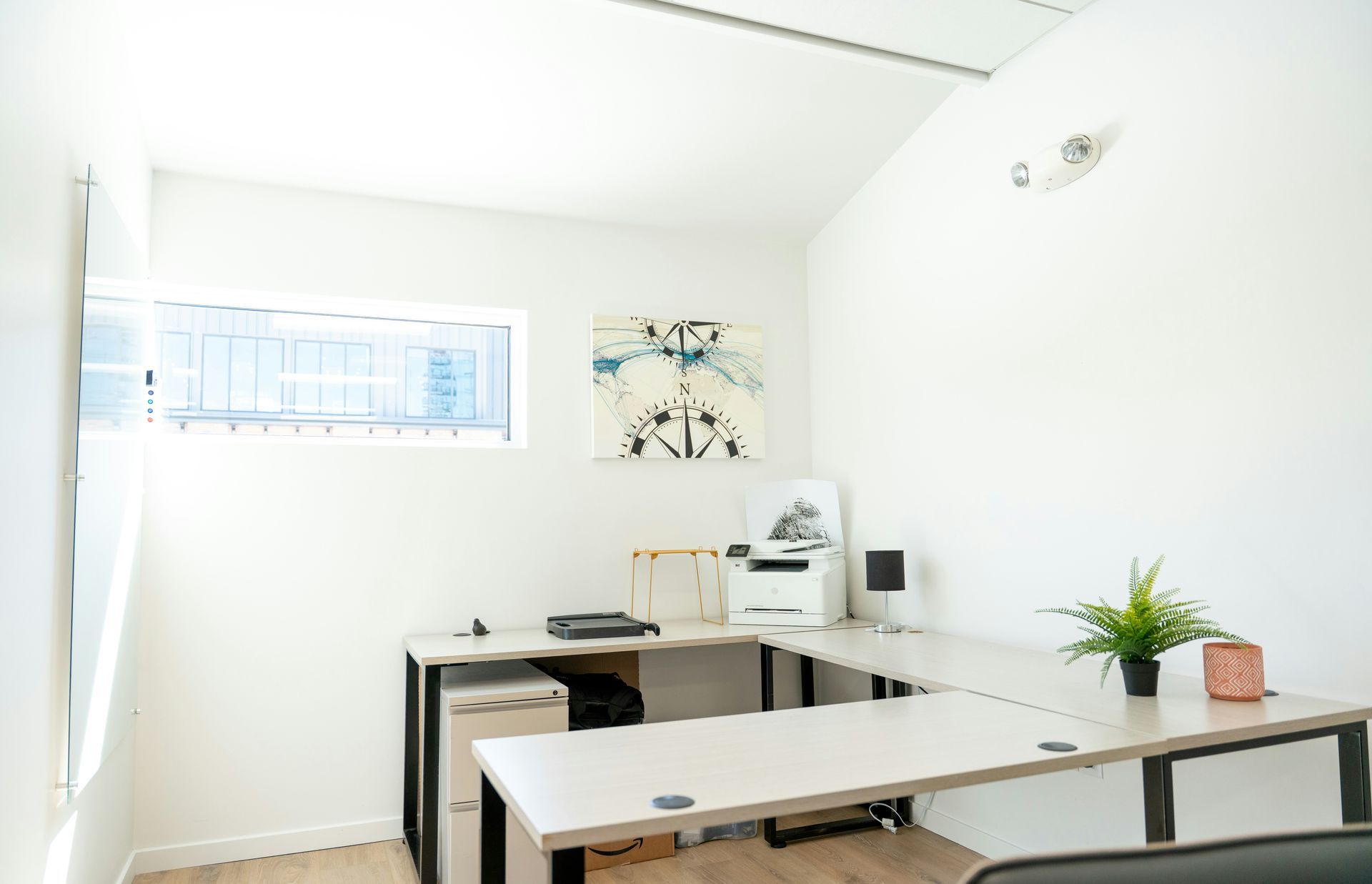 Bright office with L-shaped desk, printer, small plant, and abstract art on the wall.
