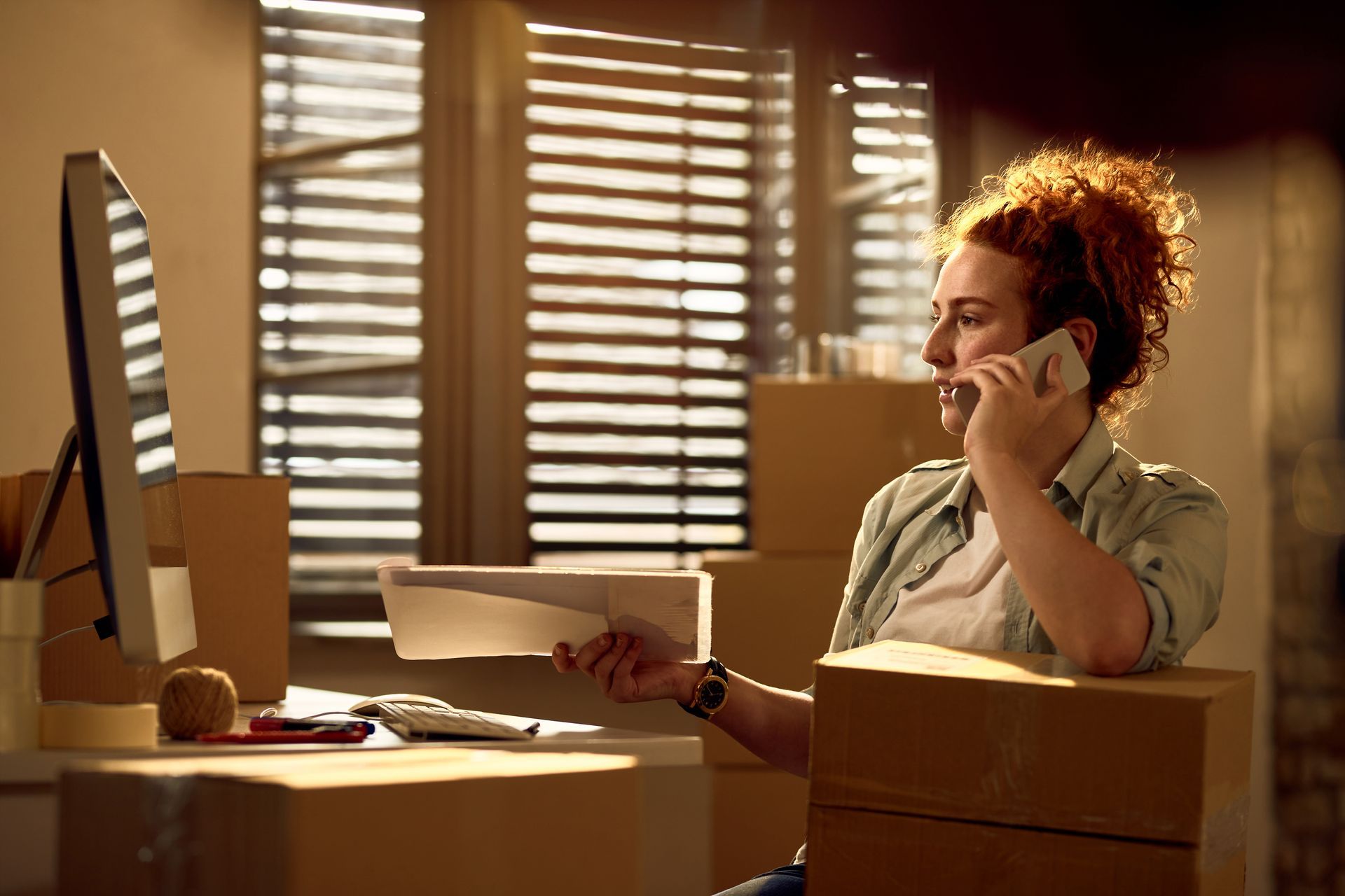 Woman with red hair on phone, working at desk with computer, packages, and paperwork near a window.