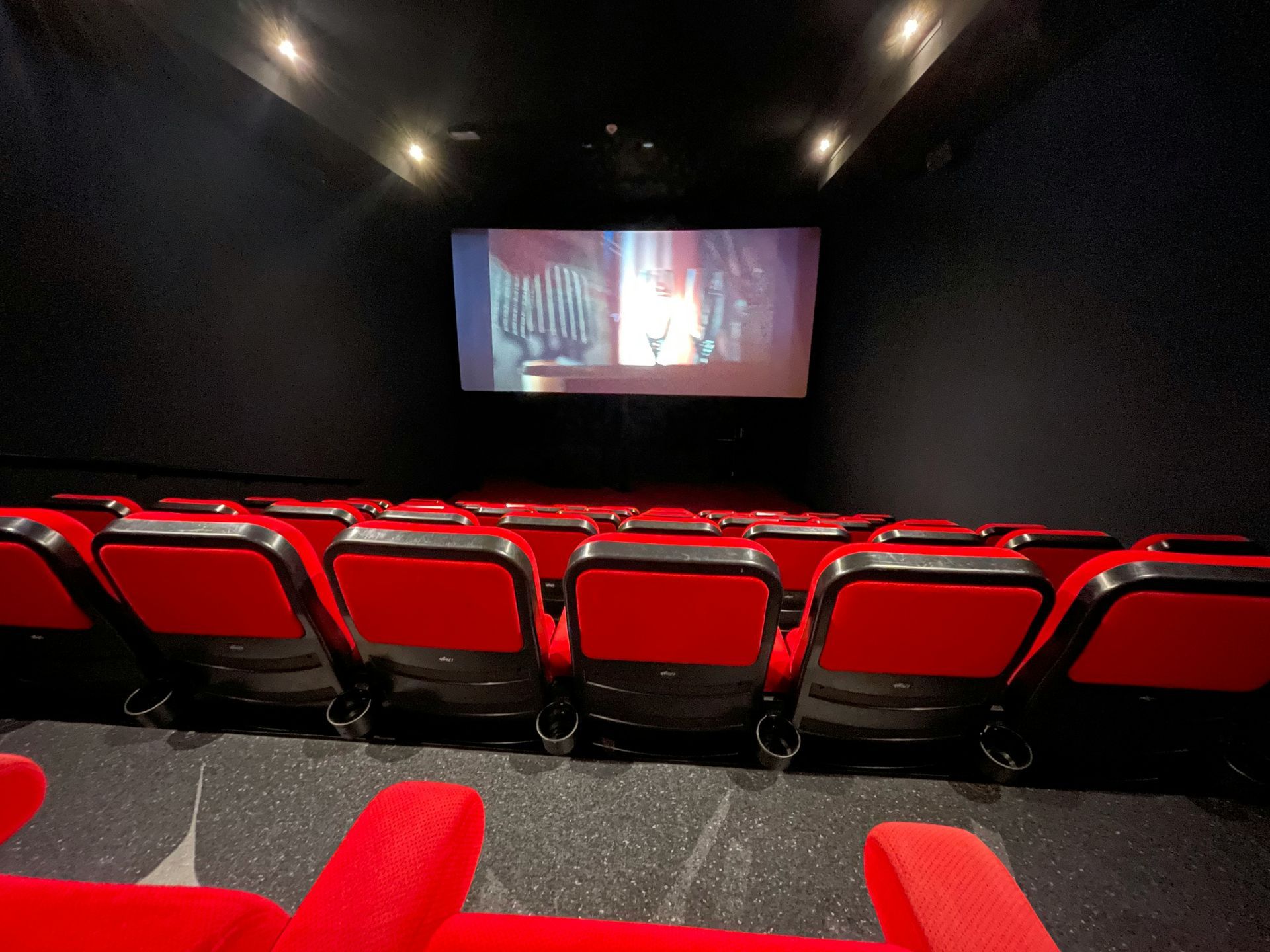 Empty movie theater with red seats and a movie playing on the screen. Dark walls and ceiling.