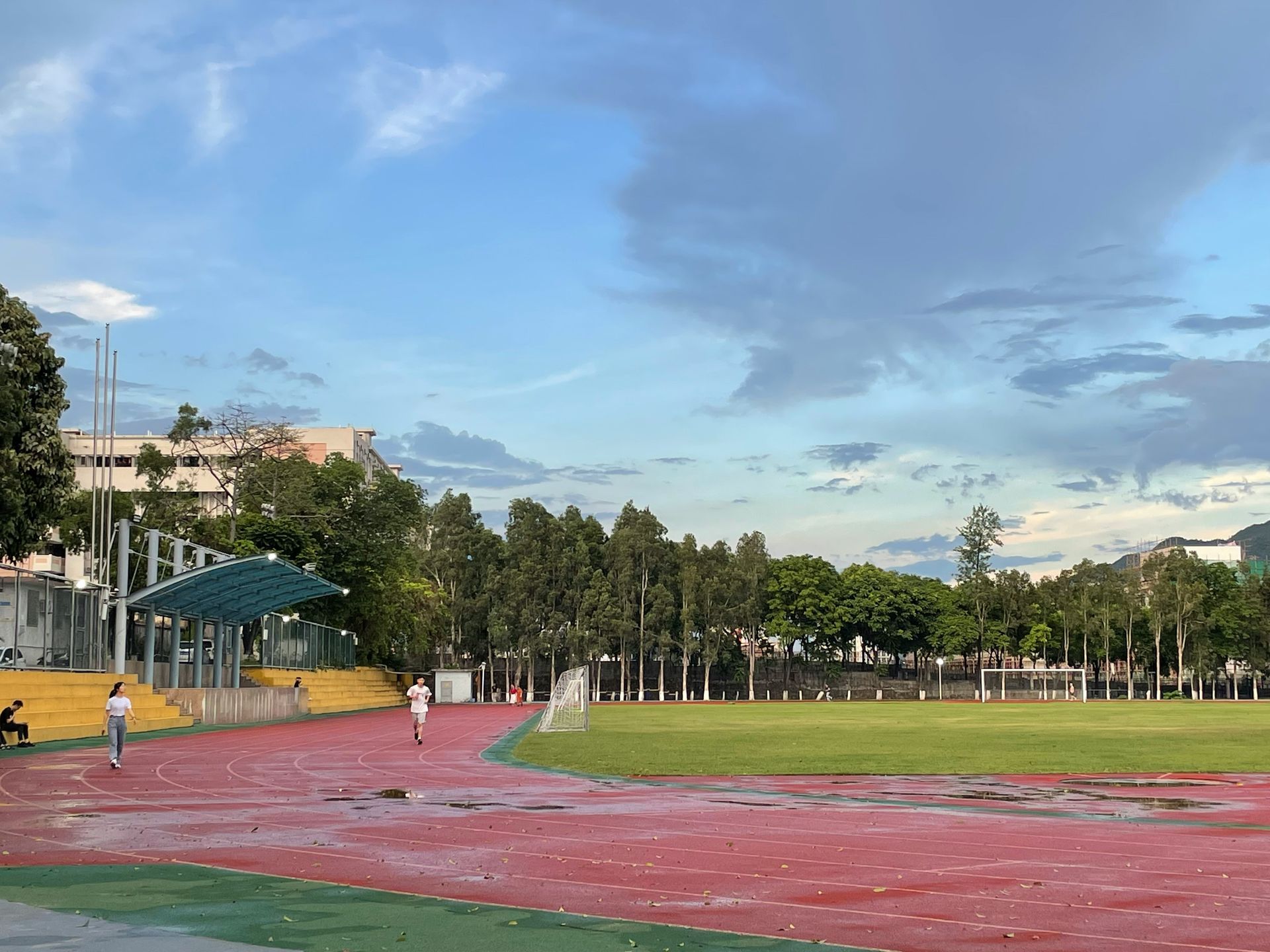 Red track around a green field at dusk, with a few people running and trees under a cloudy sky.