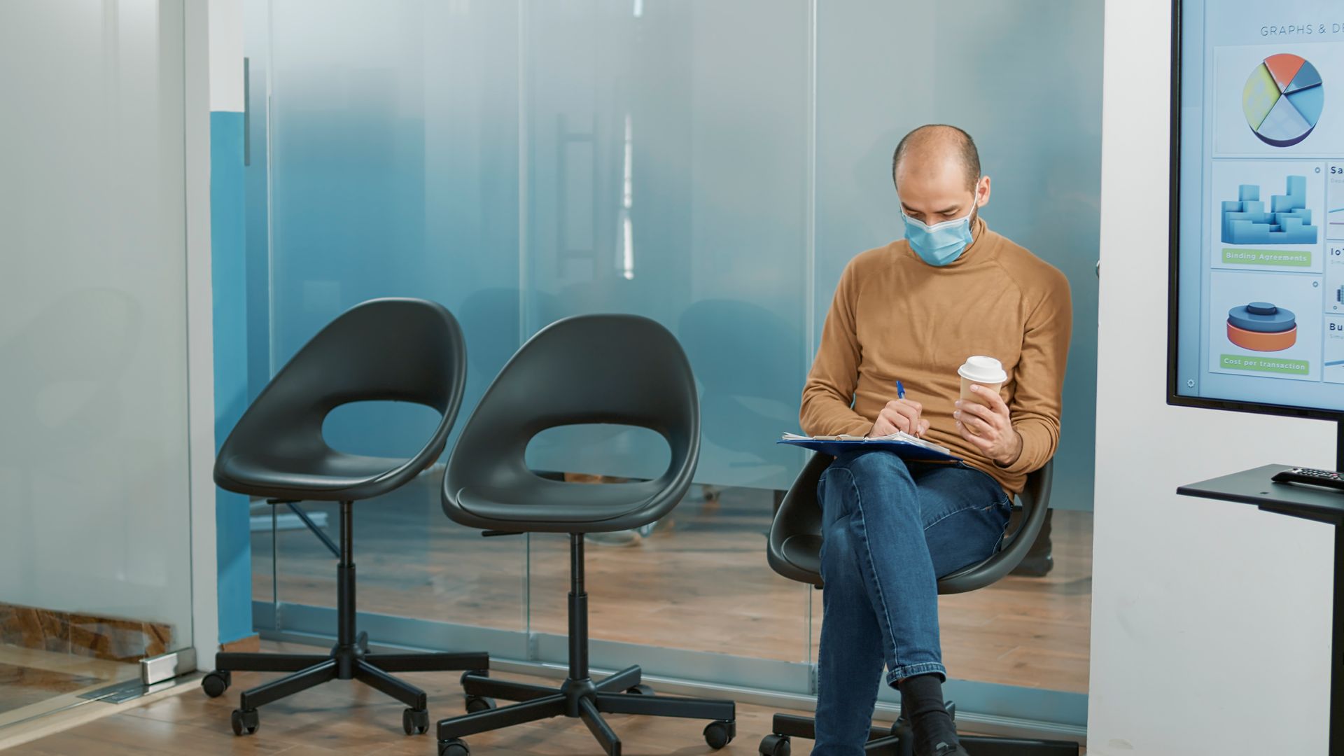 Man in Meridian wearing a mask writing in a notebook, holding a coffee cup, sitting in a waiting area.