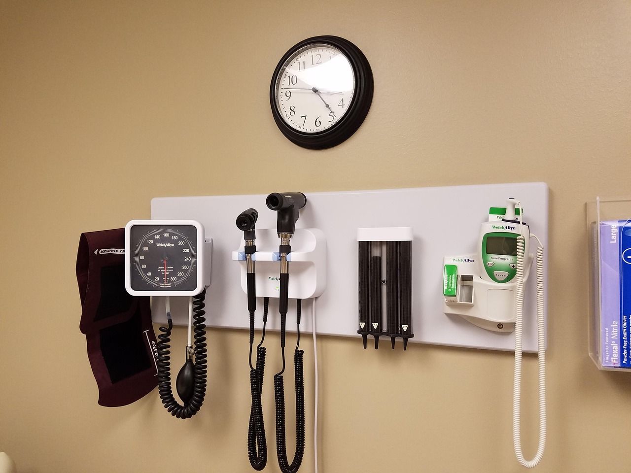 Medical exam equipment mounted on a wall, including a blood pressure gauge, clock, and otoscope.