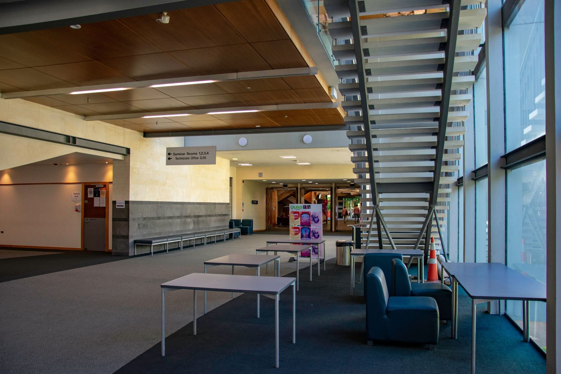 Interior of a modern building with stairs, tables, and seating area, and natural light.