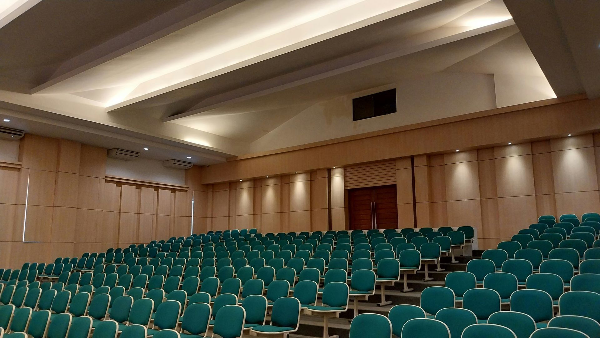 Auditorium with rows of green seating, stage, and beige walls.