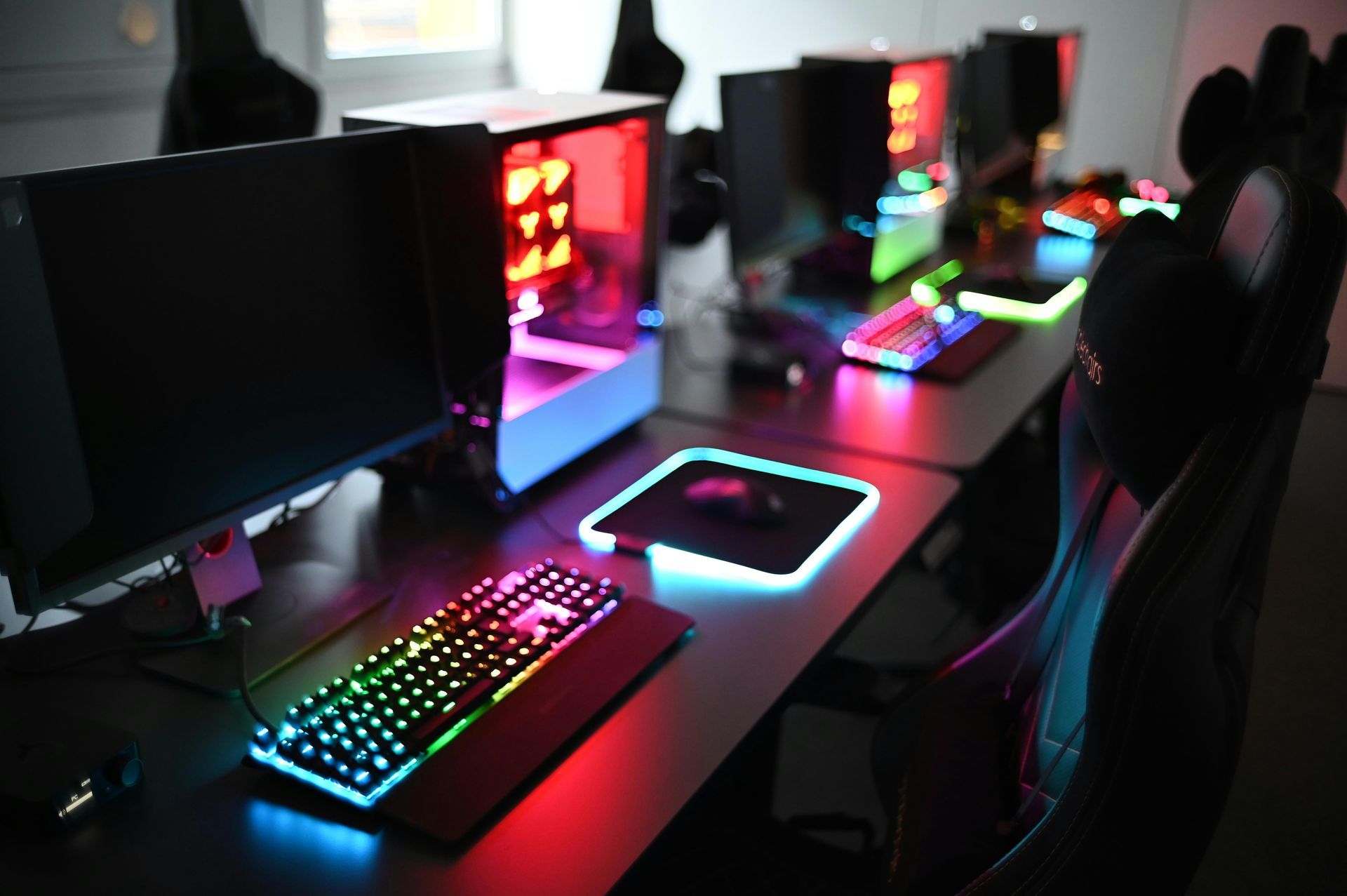 Gaming setup with colorful keyboards, monitors, and PC cases, lit up in a row.