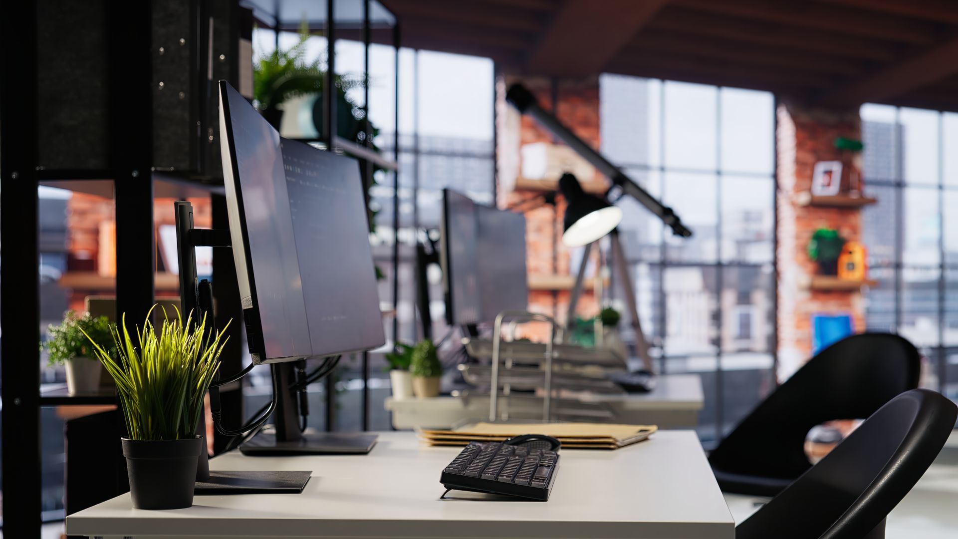 Ban Office desk in Meridian with dual monitors, keyboard, and potted plants. City view in background.