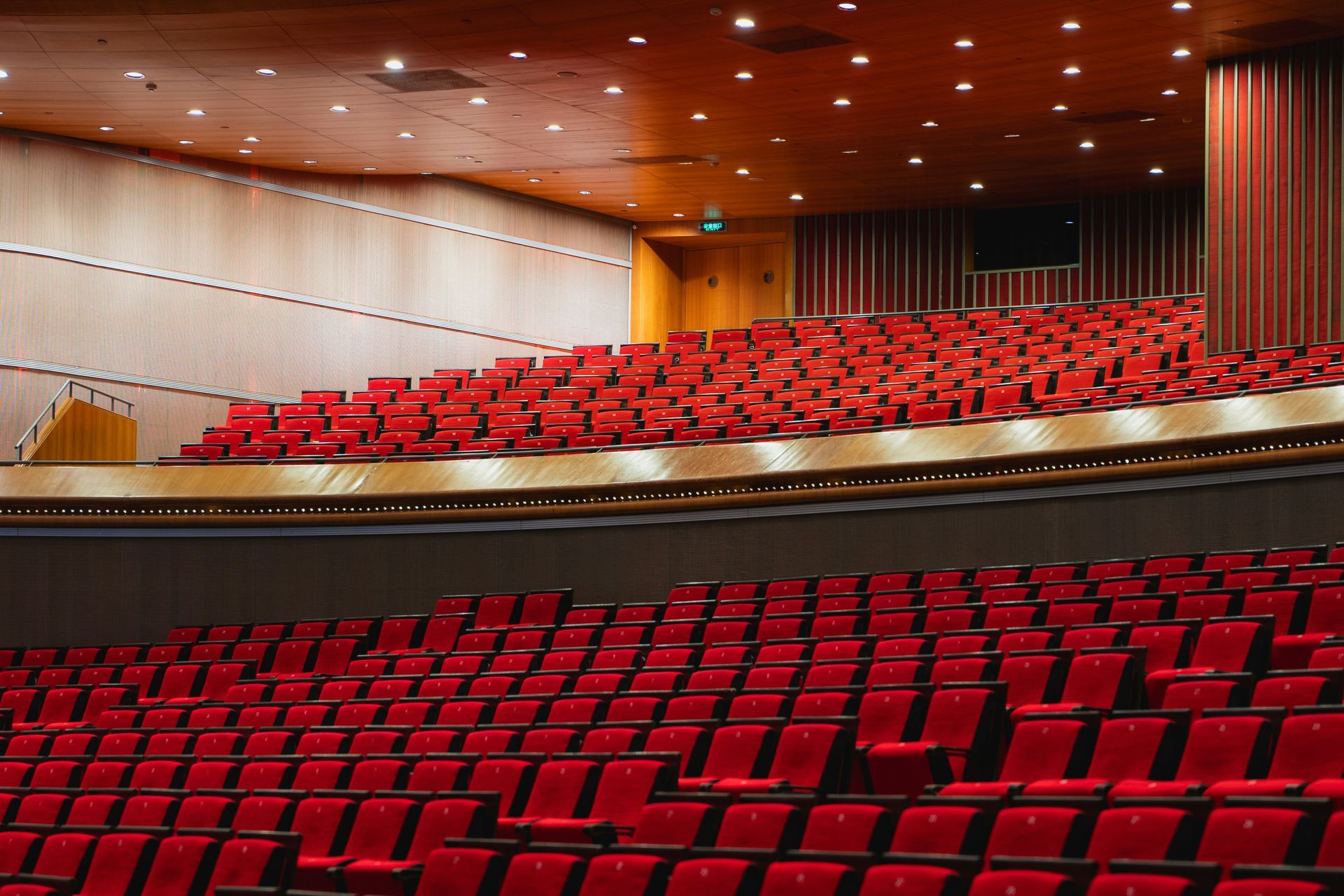 Empty red theater seats, tiered, facing a stage. Brown wood paneling on walls and ceiling.