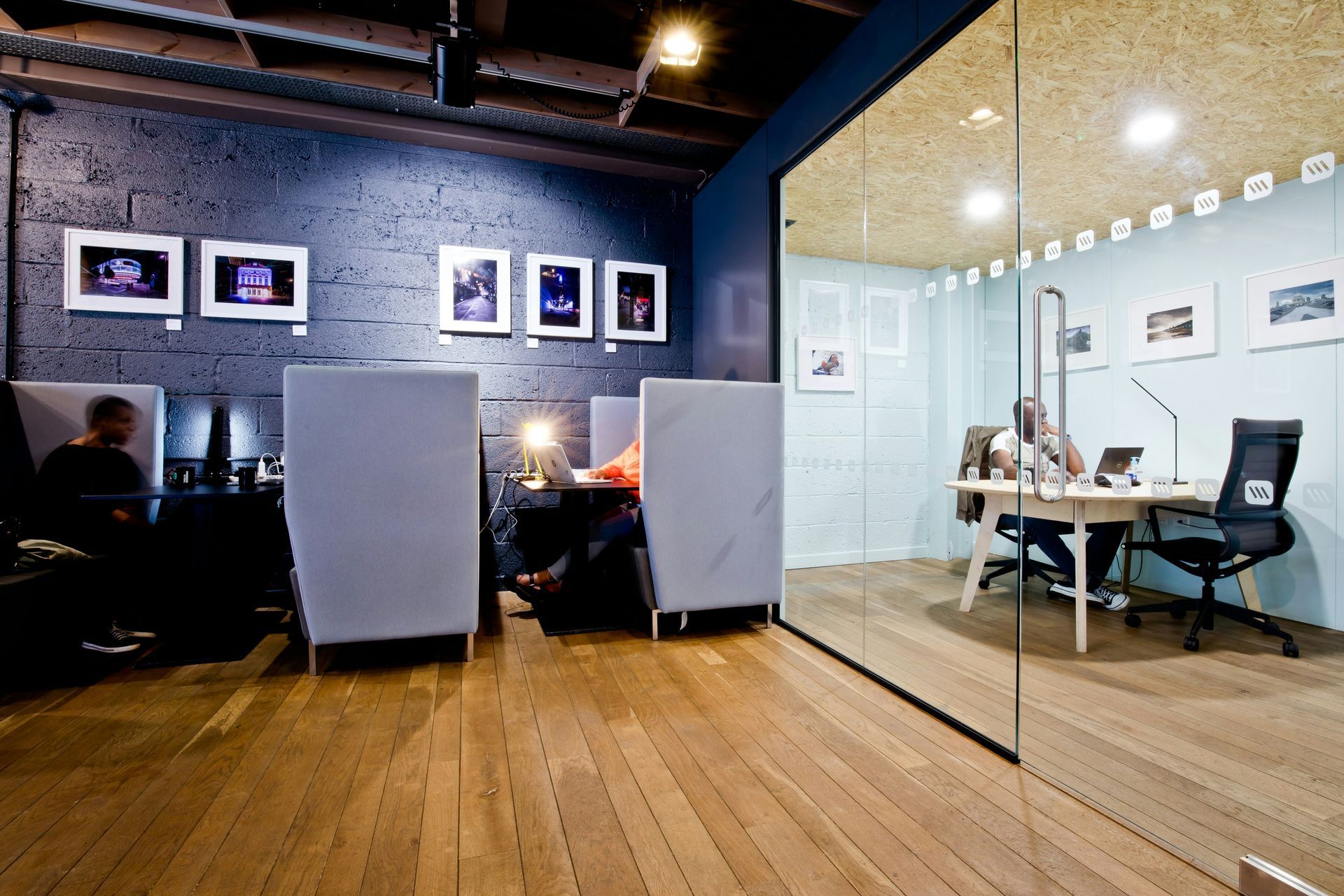 A modern office space with wood floors, a glass-walled room, and individuals working at desks.
