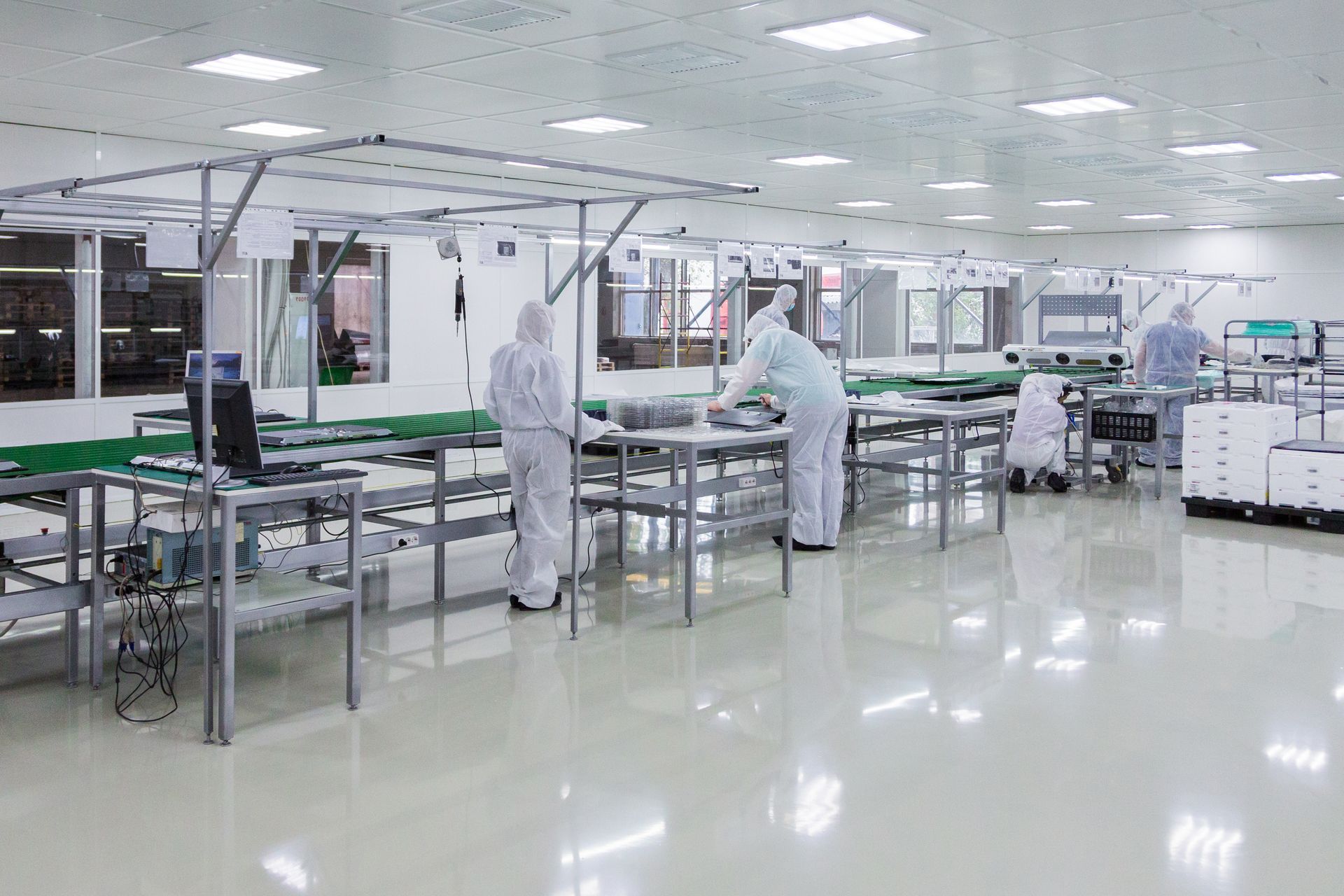 People in clean suits working in a bright, sterile factory. Workstations and overhead support structure visible.